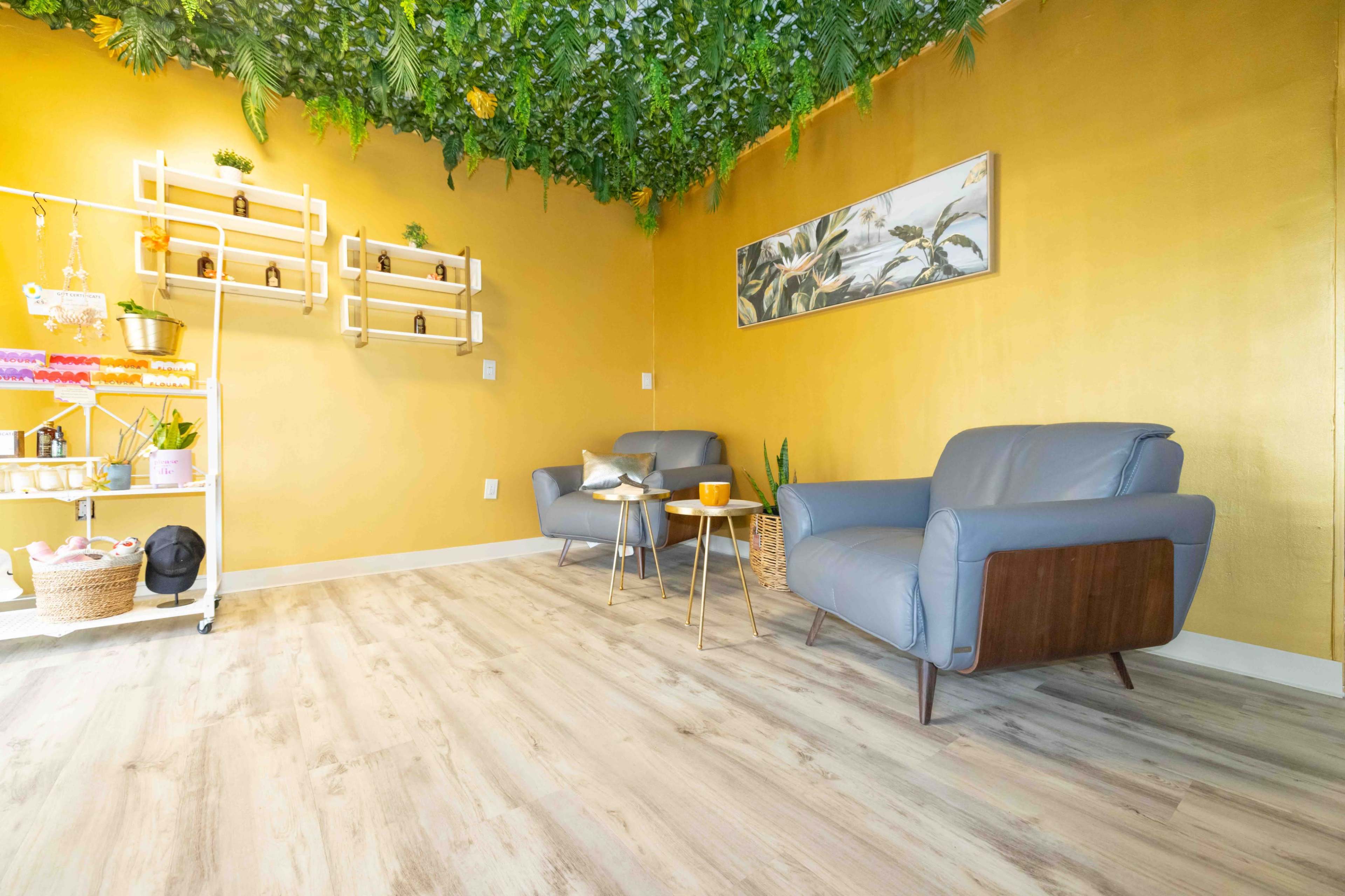 The image shows a cozy room with two blue armchairs, a small side table, a wall decorated with greenery, and shelves displaying various items against a golden wall.