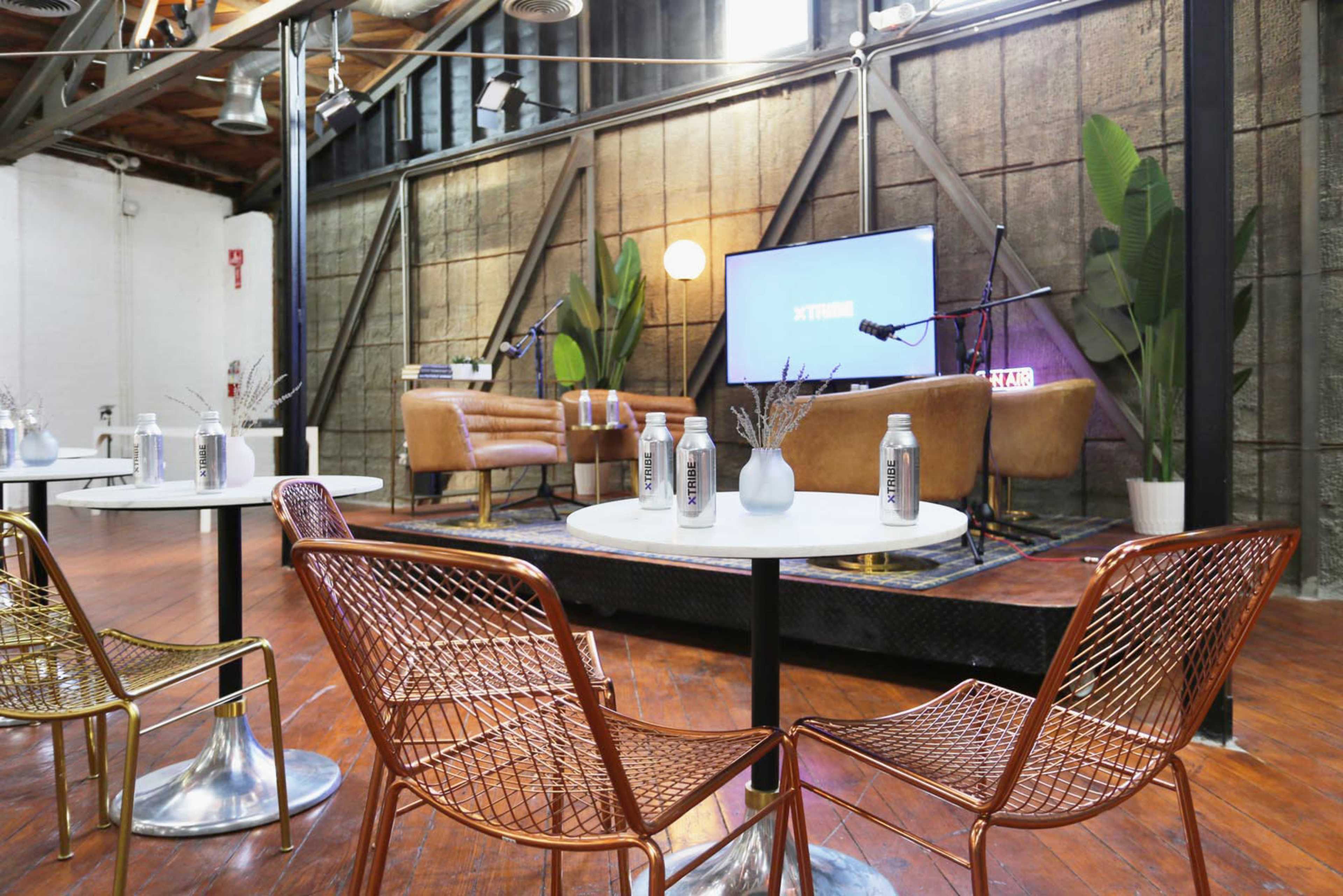 A styled event space featuring round tables with metallic chairs, a pair of brown leather sofas, and a large screen against a textured wall, complemented by plants and microphones.