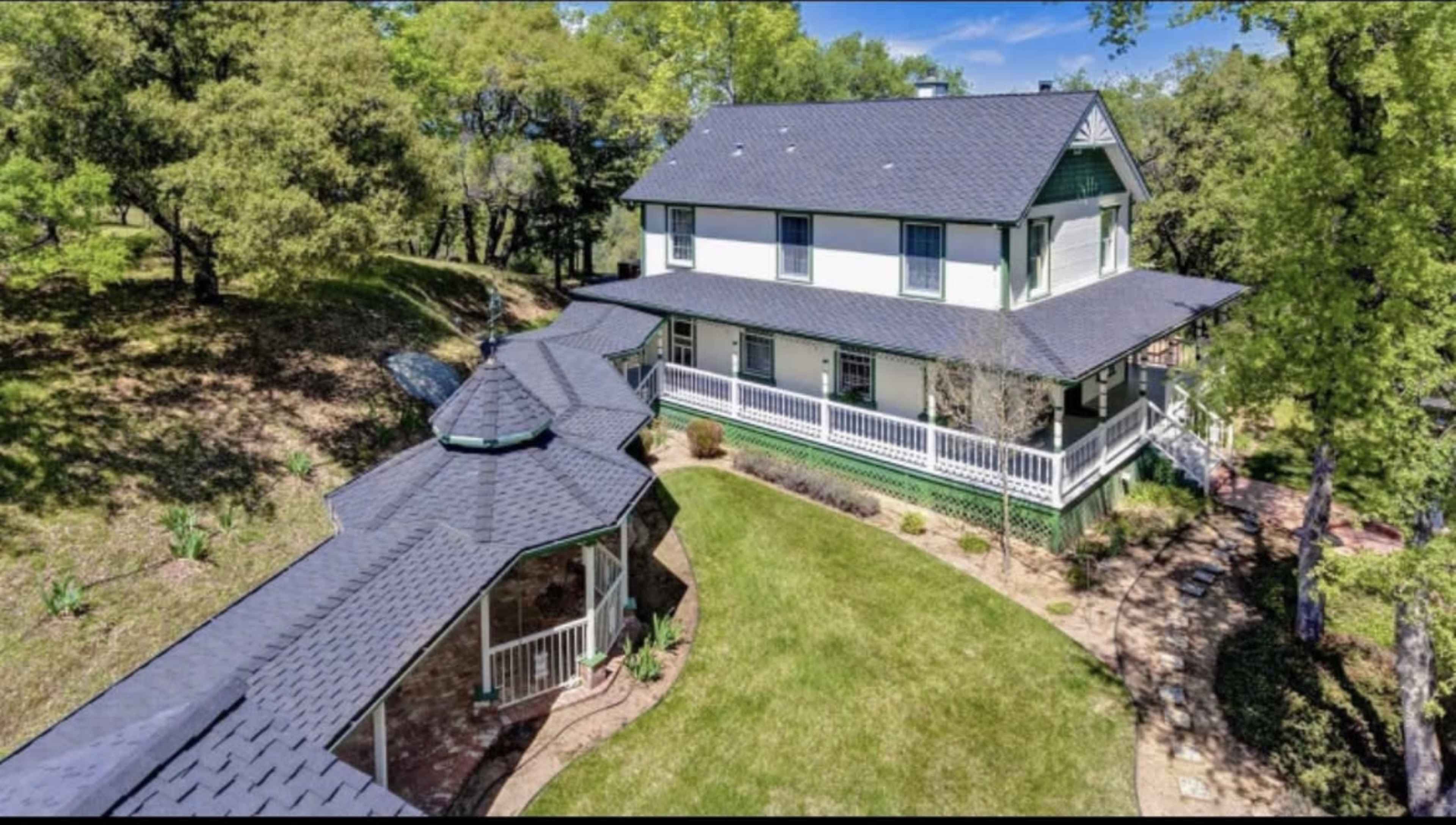 A two-story house with a large porch and a curved pathway is surrounded by trees and greenery.
