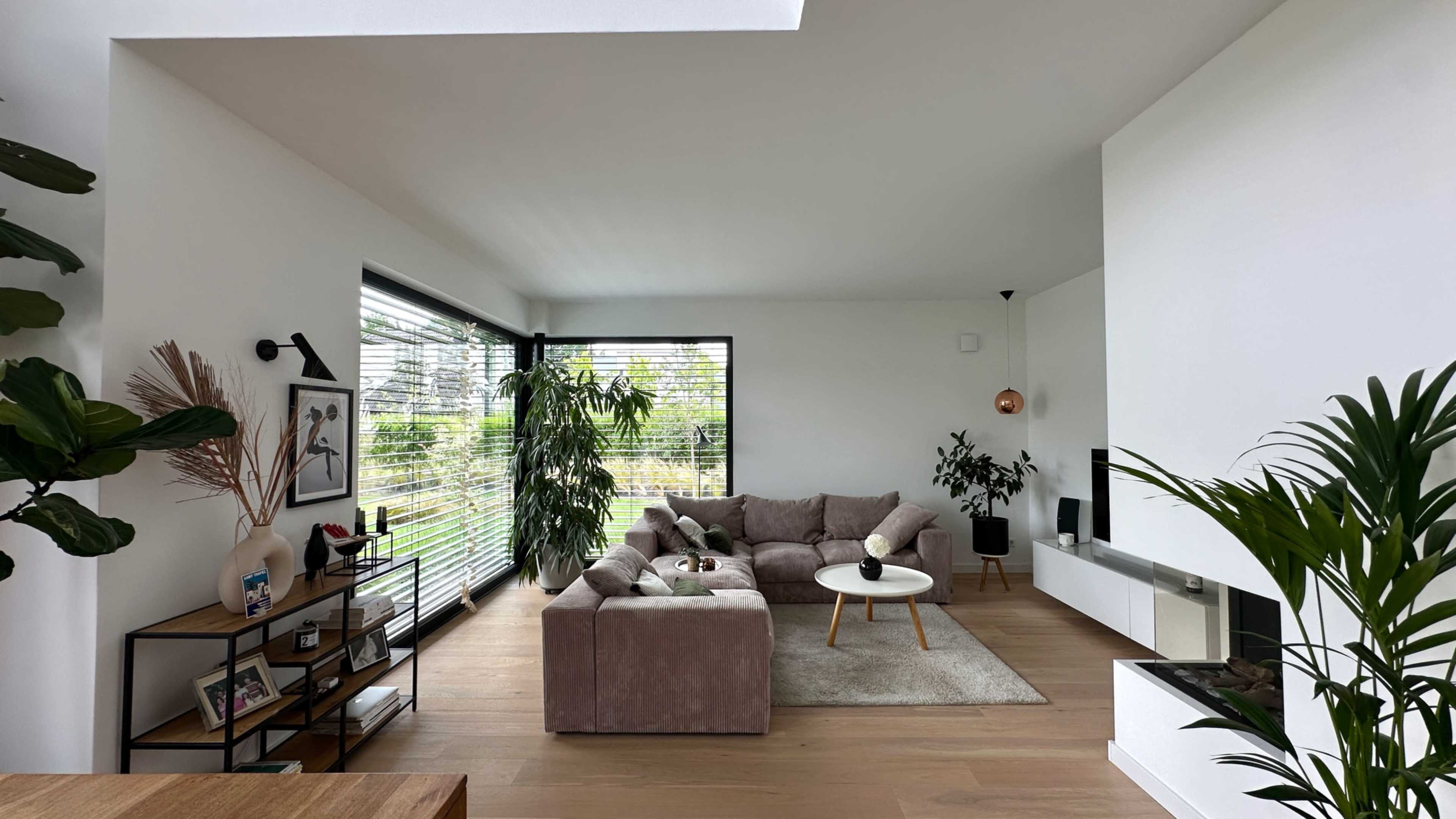The image shows a modern living room featuring a light-colored sofa, a round coffee table, and large windows with greenery outside.