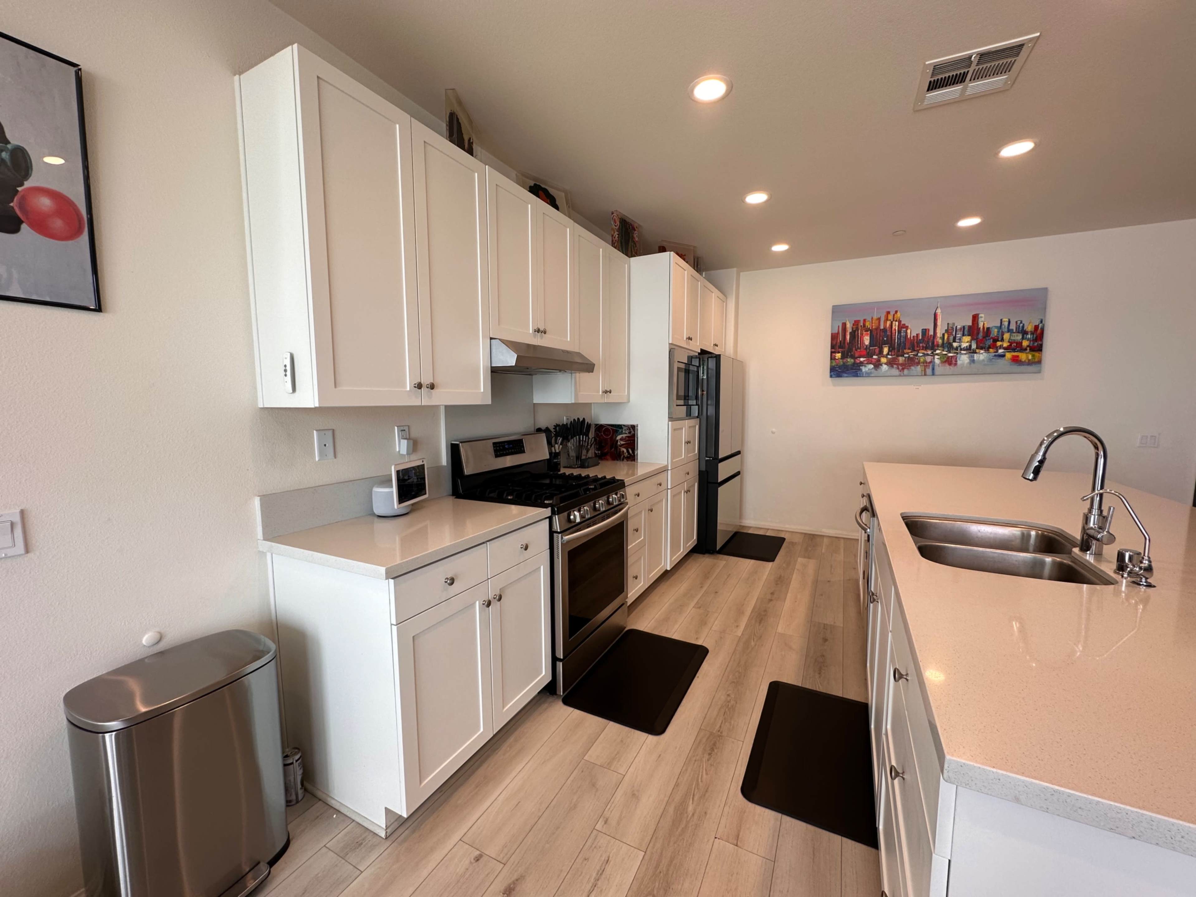 A modern kitchen features white cabinets, stainless steel appliances, a countertop with a sink, and a cityscape painting on the wall.