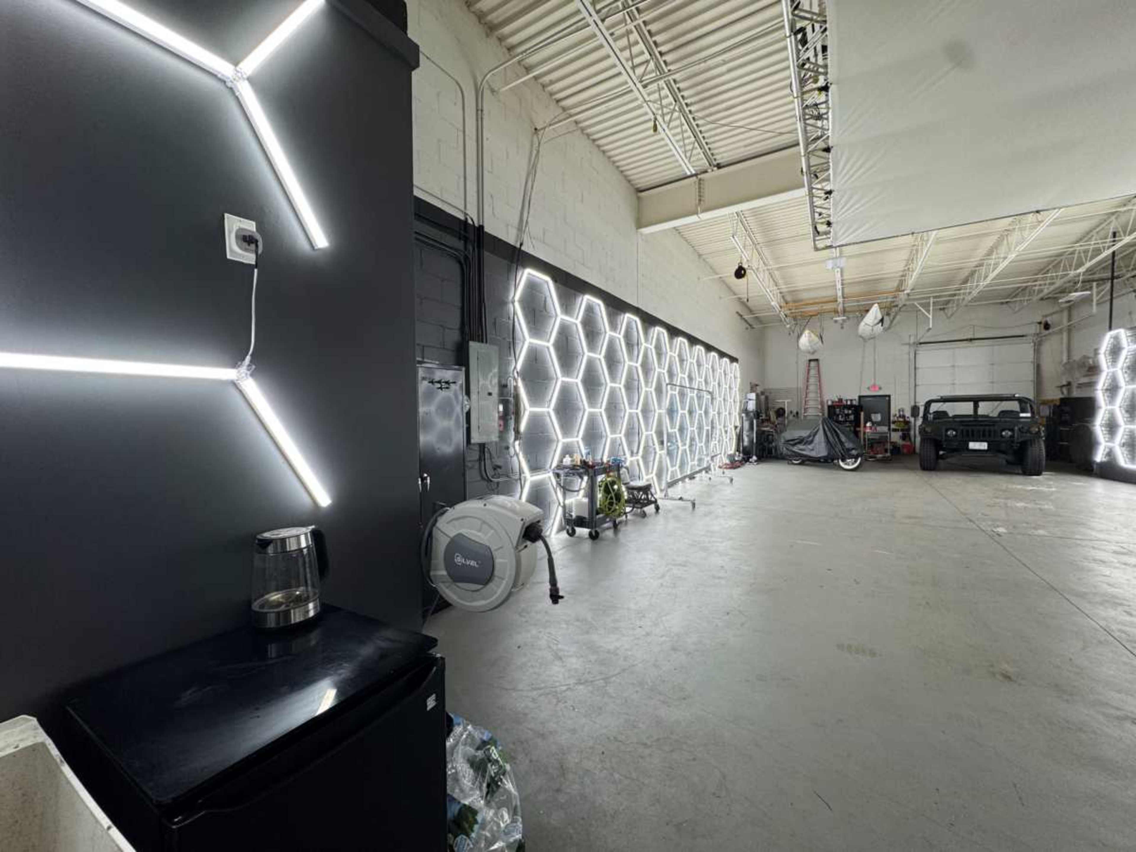 The image shows a spacious industrial interior with hexagonal light fixtures lining one wall and a black refrigerator in the foreground.