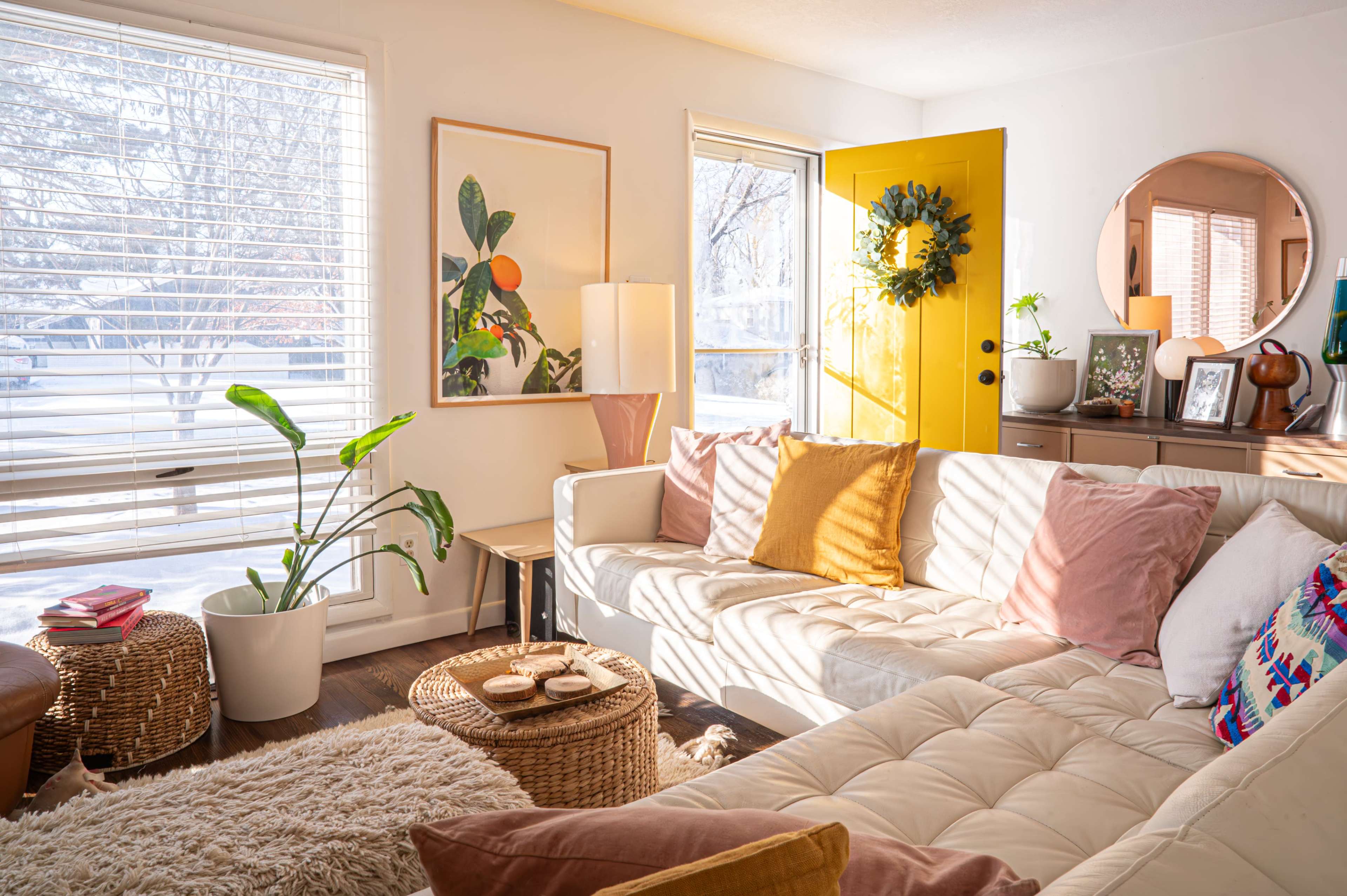 The image showcases a well-lit living room featuring a white sofa with colorful cushions, a round mirror, and a yellow front door.