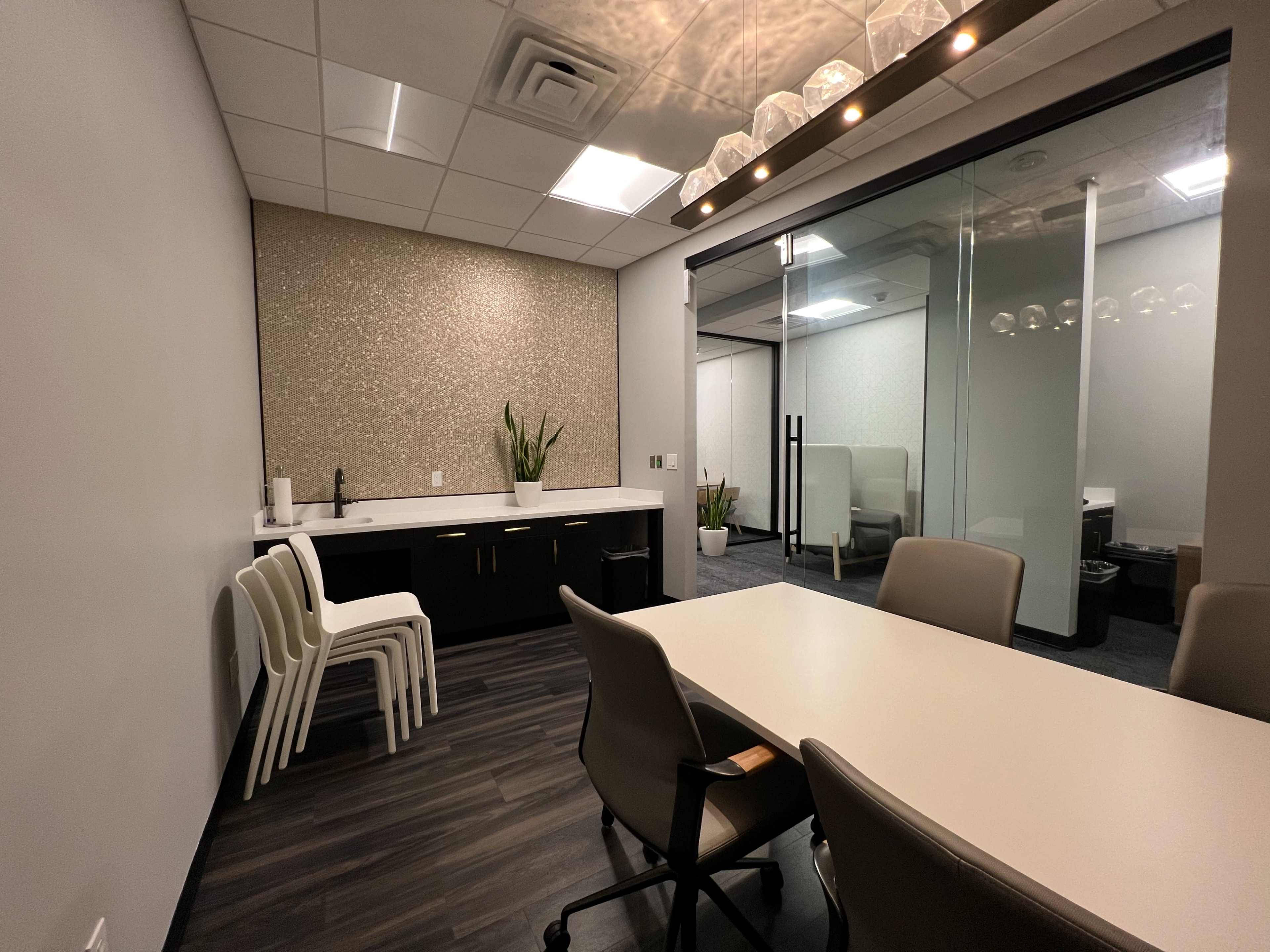 A modern conference room with a table, office chairs, and a kitchenette area, featuring a glass partition leading to another workspace.