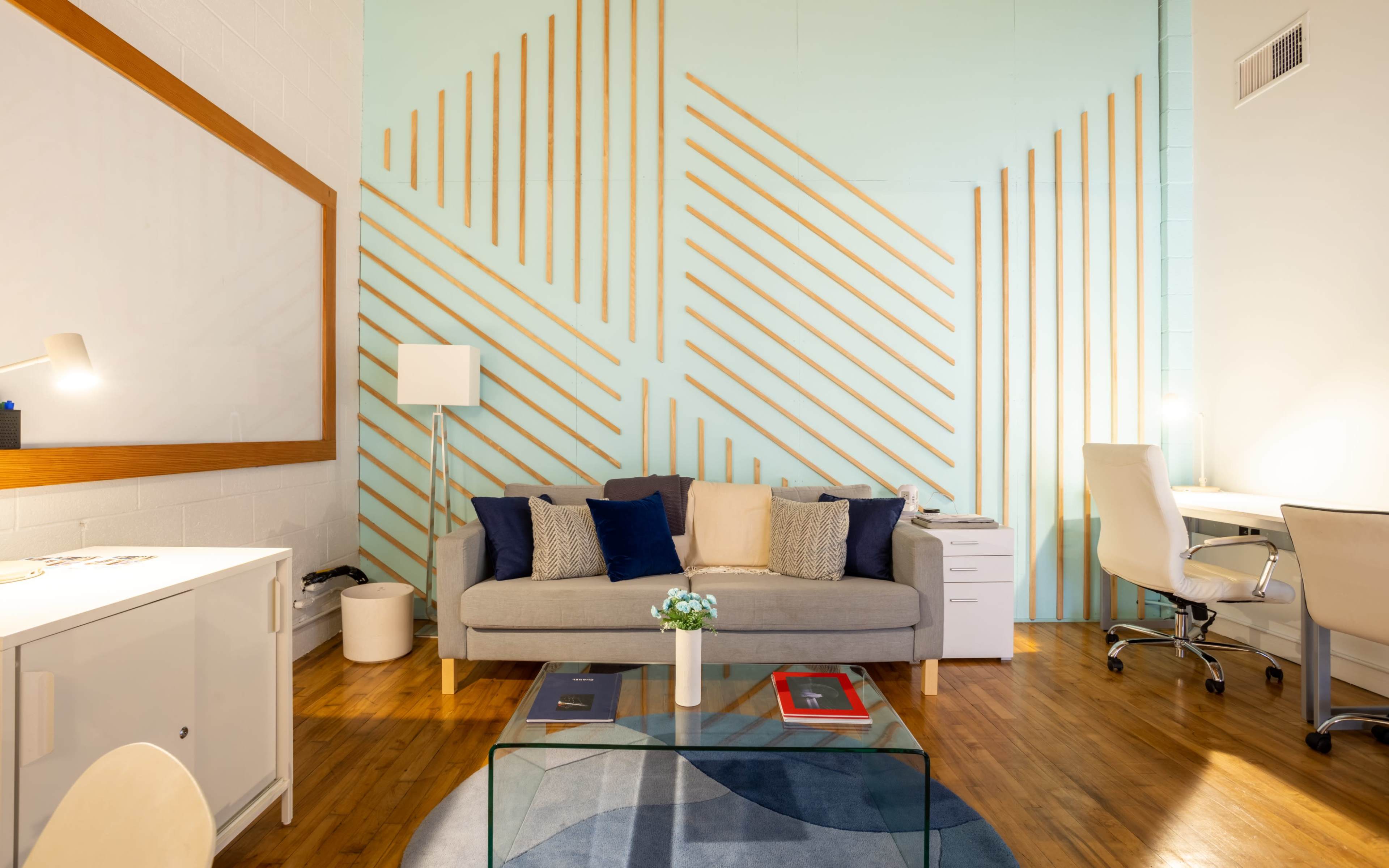 The image shows a modern living space featuring a light blue wall with wooden lines, a gray sofa with cushions, a glass coffee table, and a desk setup in the corner.