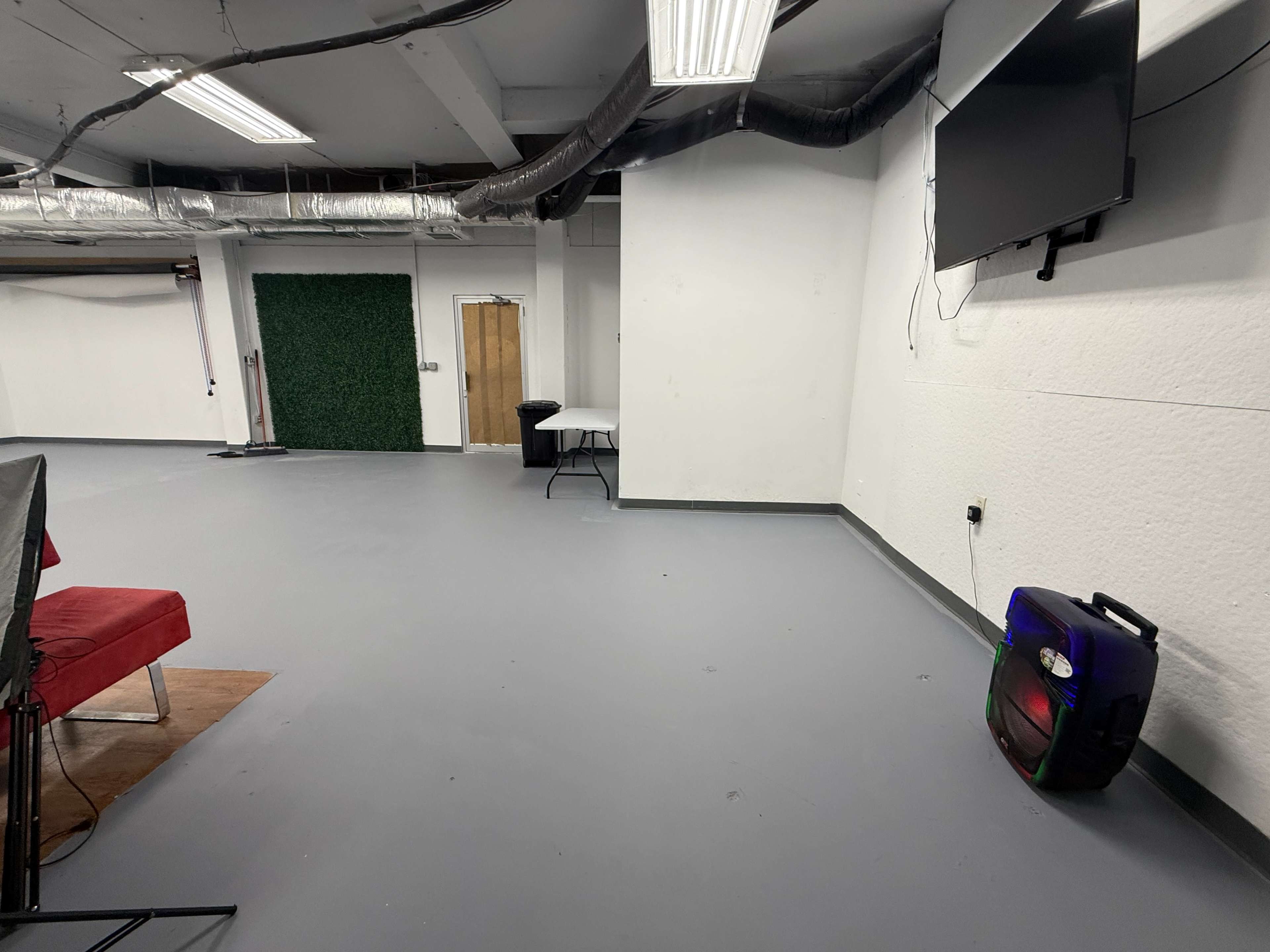 The image shows a spacious, empty room with a grey floor, white walls, a table, and a television mounted on the wall.