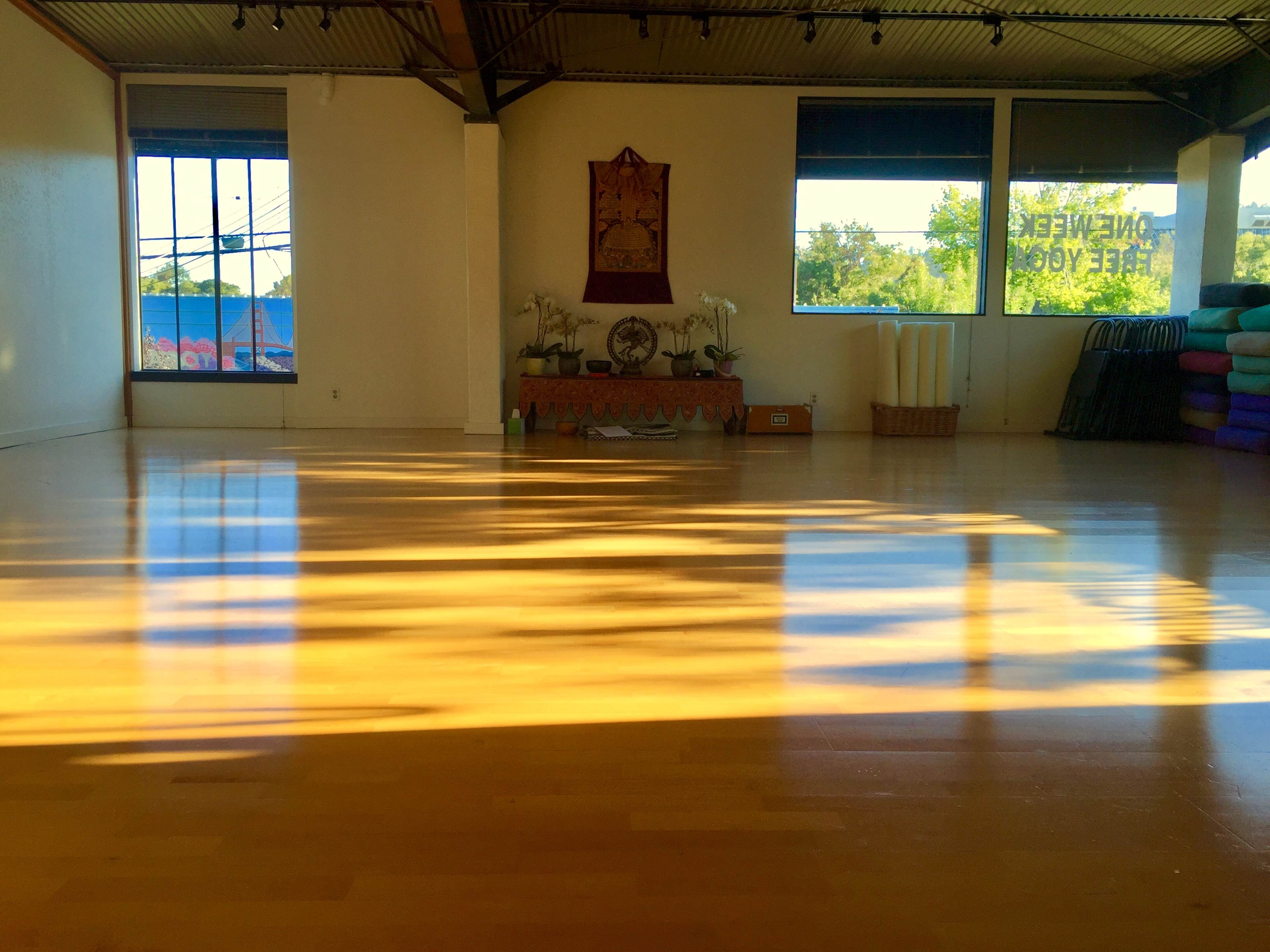A spacious yoga studio features polished wooden floors illuminated by sunlight, with yoga mats stacked against the wall and decorative elements at the front.