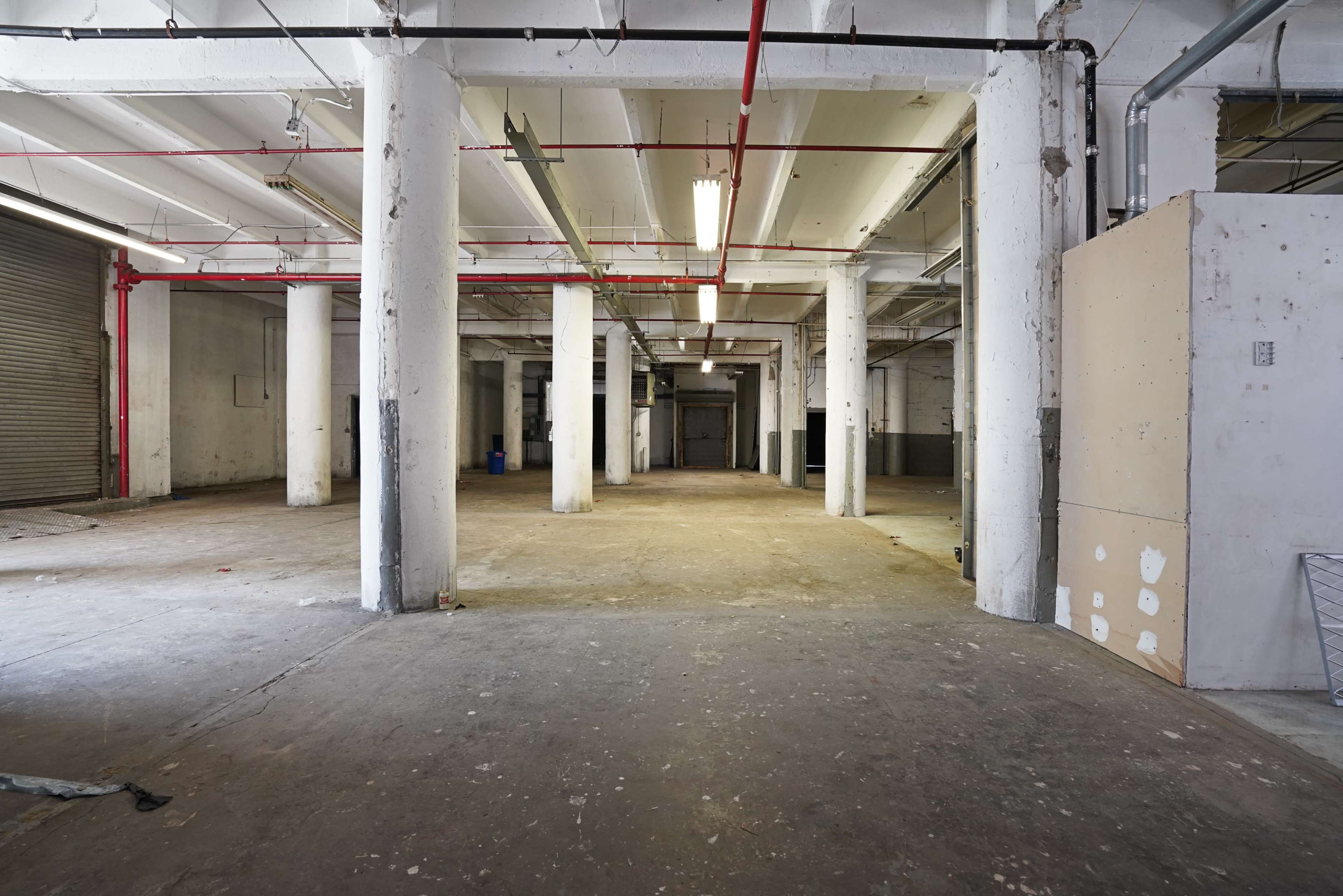 The image shows an empty industrial space with concrete floors, white columns, and exposed overhead piping.