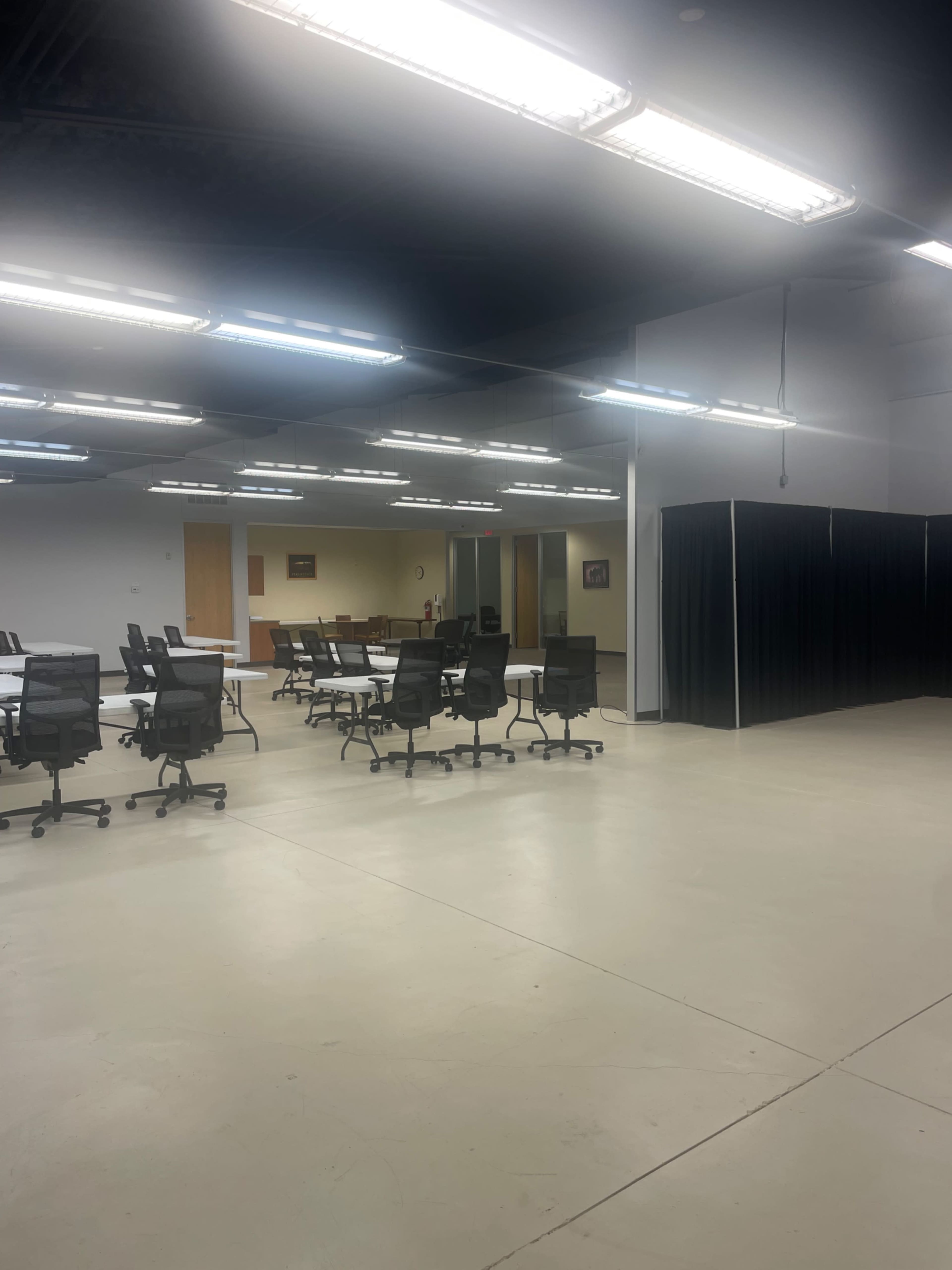 A spacious room with several black rolling chairs arranged around tables, illuminated by overhead fluorescent lights, and featuring a black curtain in one corner.