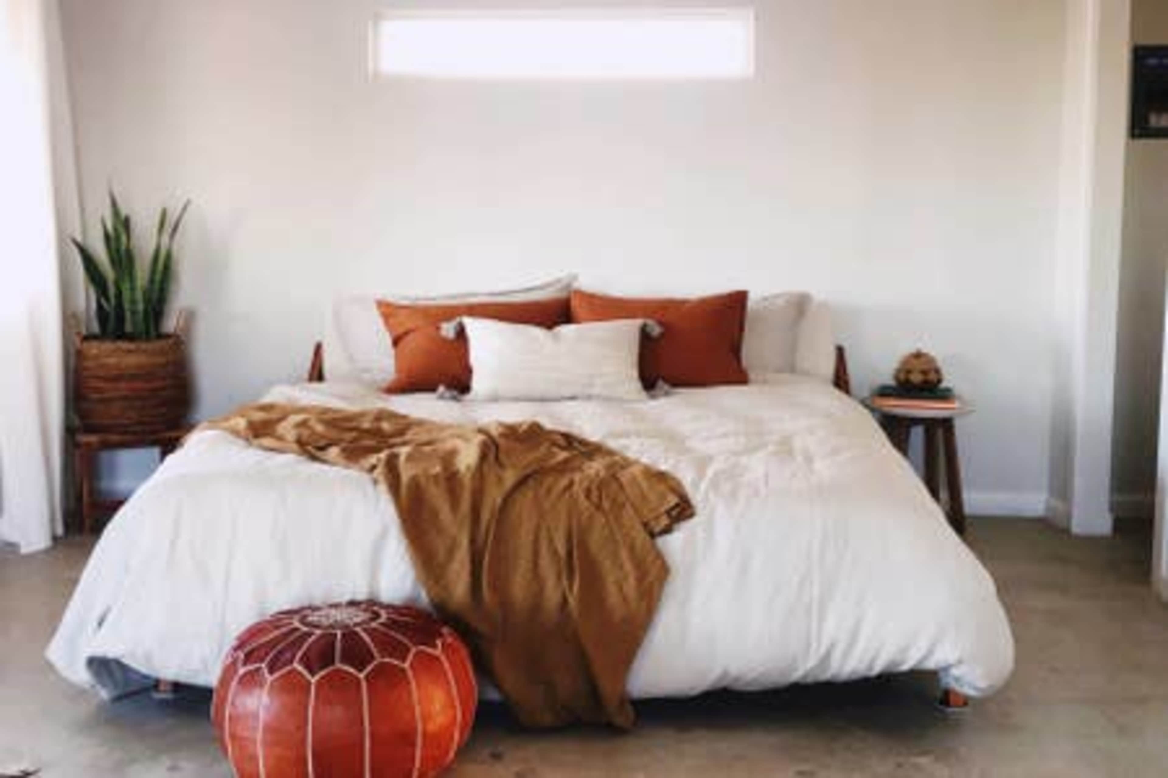 A neatly arranged bedroom features a large bed with white linens and several pillows, accompanied by a brown throw blanket and a decorative pouf.