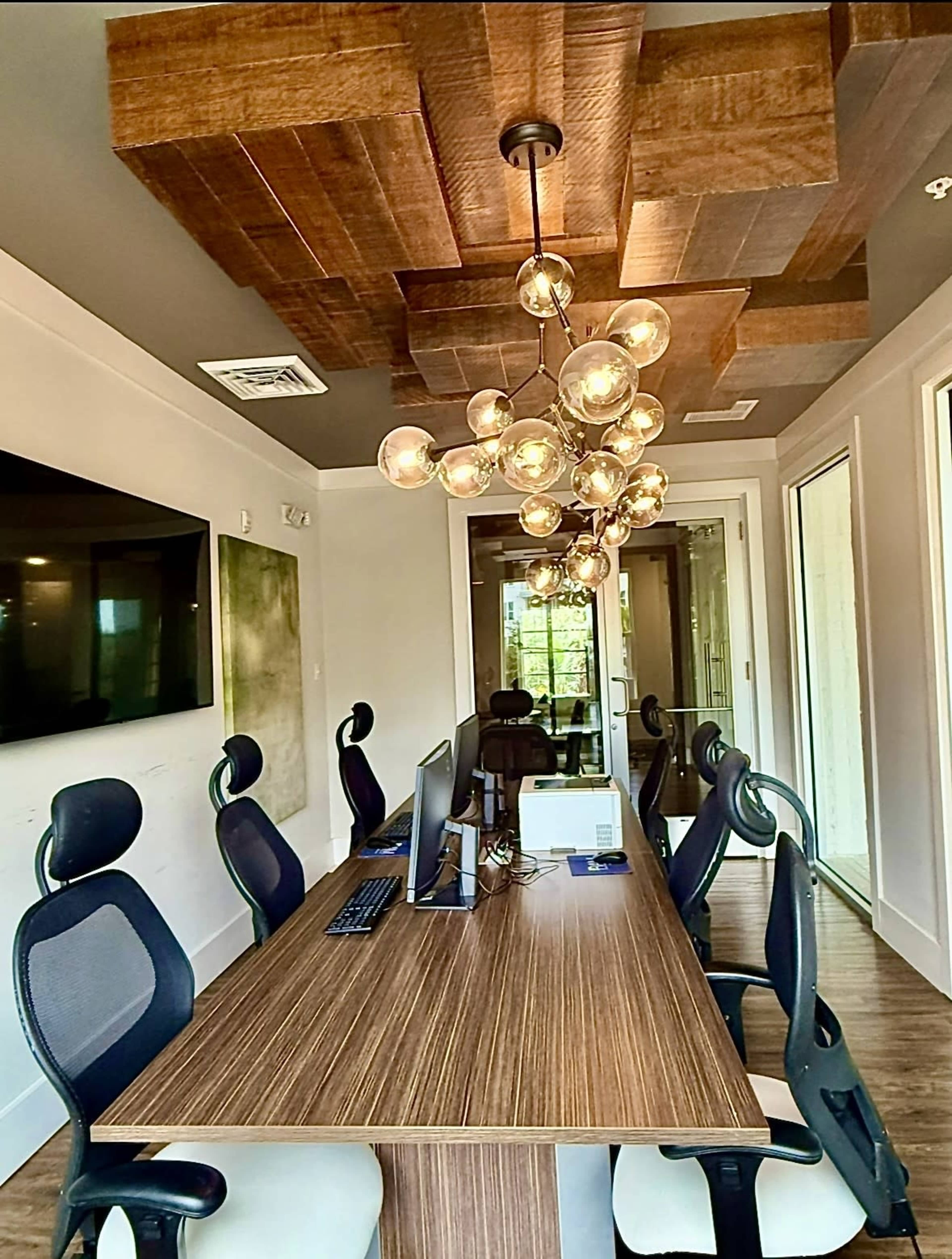 The image shows a modern office space featuring a large wooden meeting table, ergonomic chairs, and a decorative chandelier above it.
