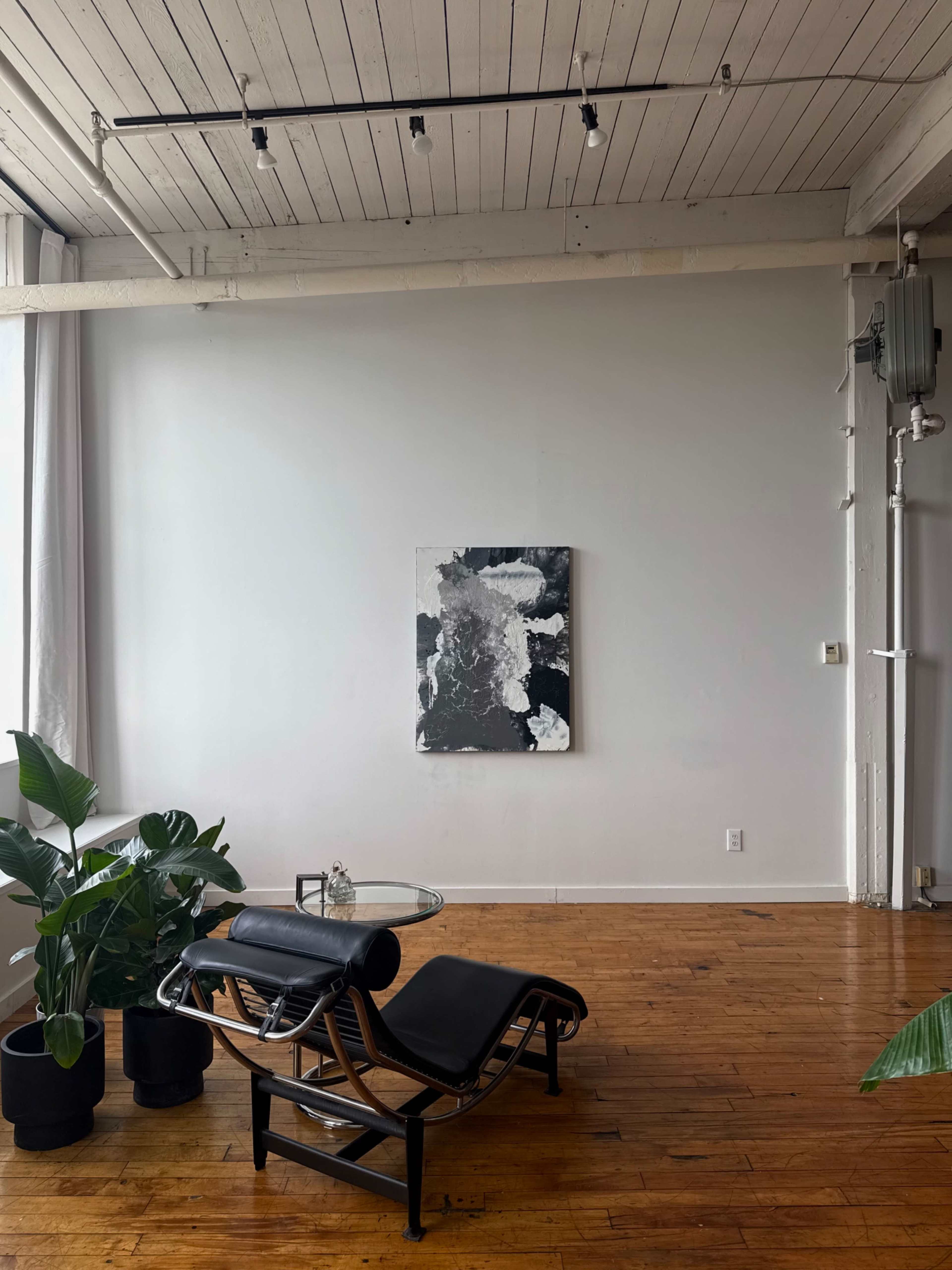 A minimalist interior features a chaise lounge and a glass table in front of a large black-and-white abstract painting on the wall.