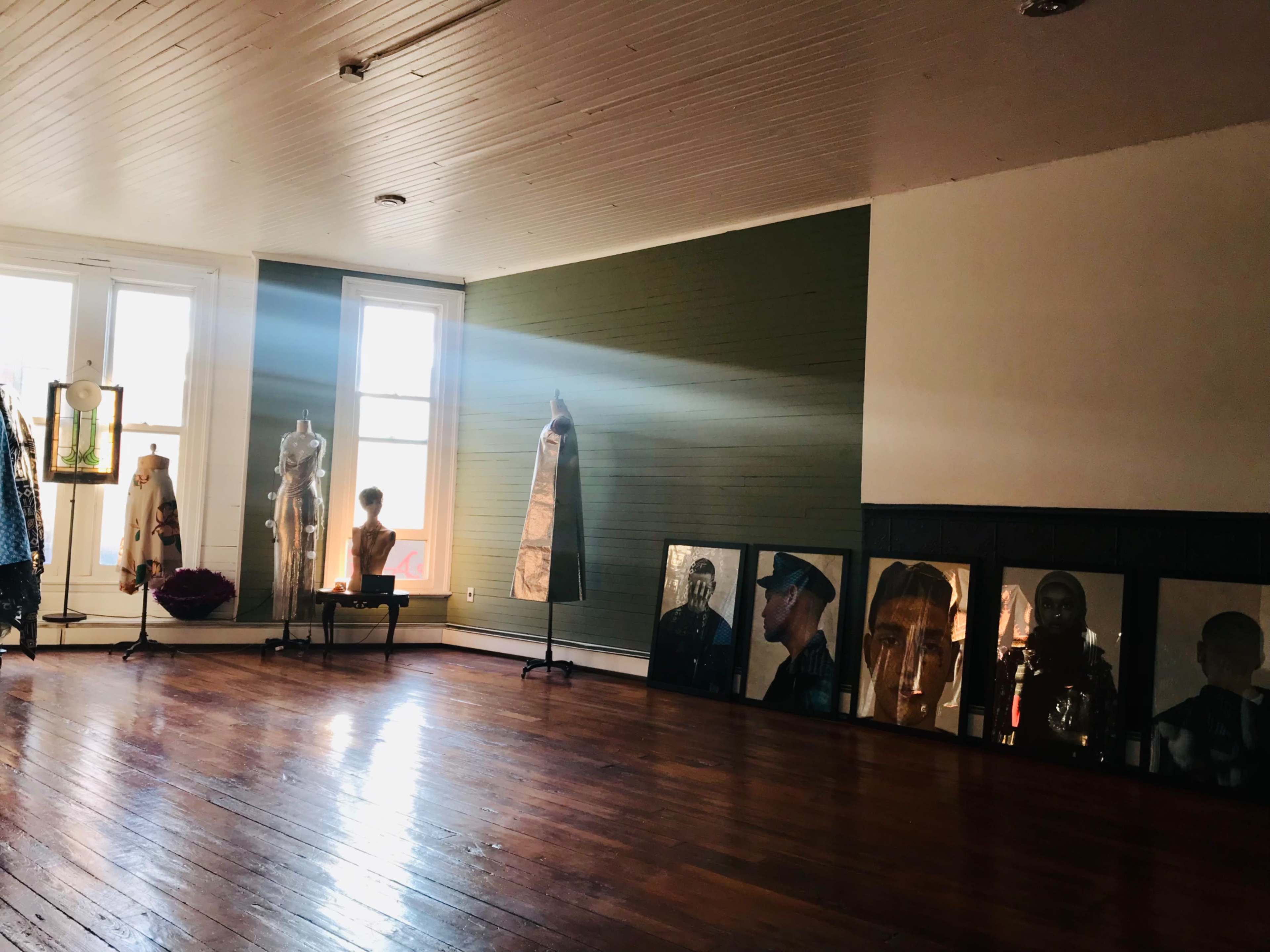 The image shows a spacious room with wooden floors, featuring mannequins dressed in clothing and framed portraits of people along one wall.