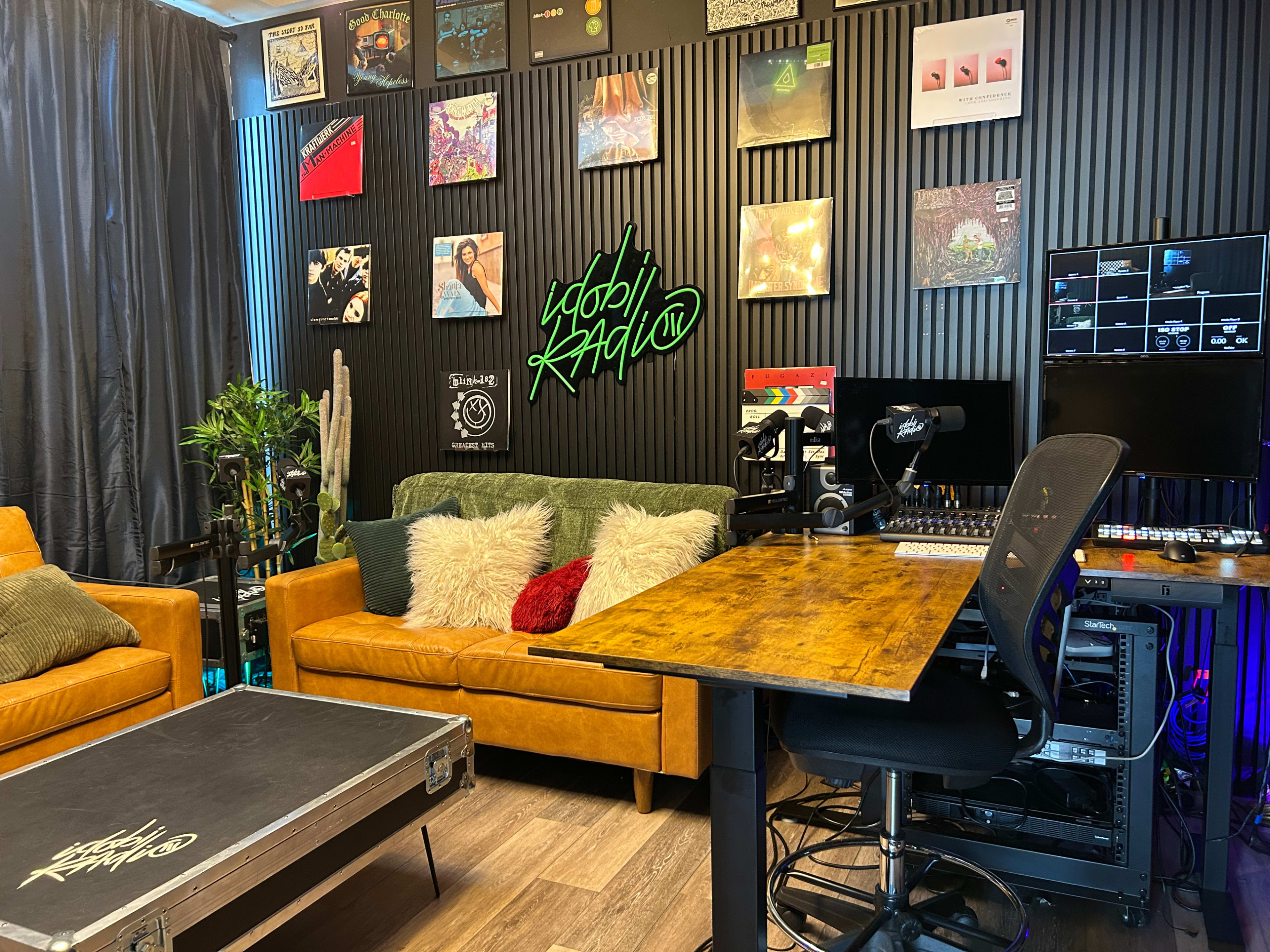 The image depicts a modern recording studio featuring a wooden desk, a black ergonomic chair, orange leather couches adorned with decorative pillows, and a wall decorated with vinyl records and a neon sign that reads "Yobbi Radio."