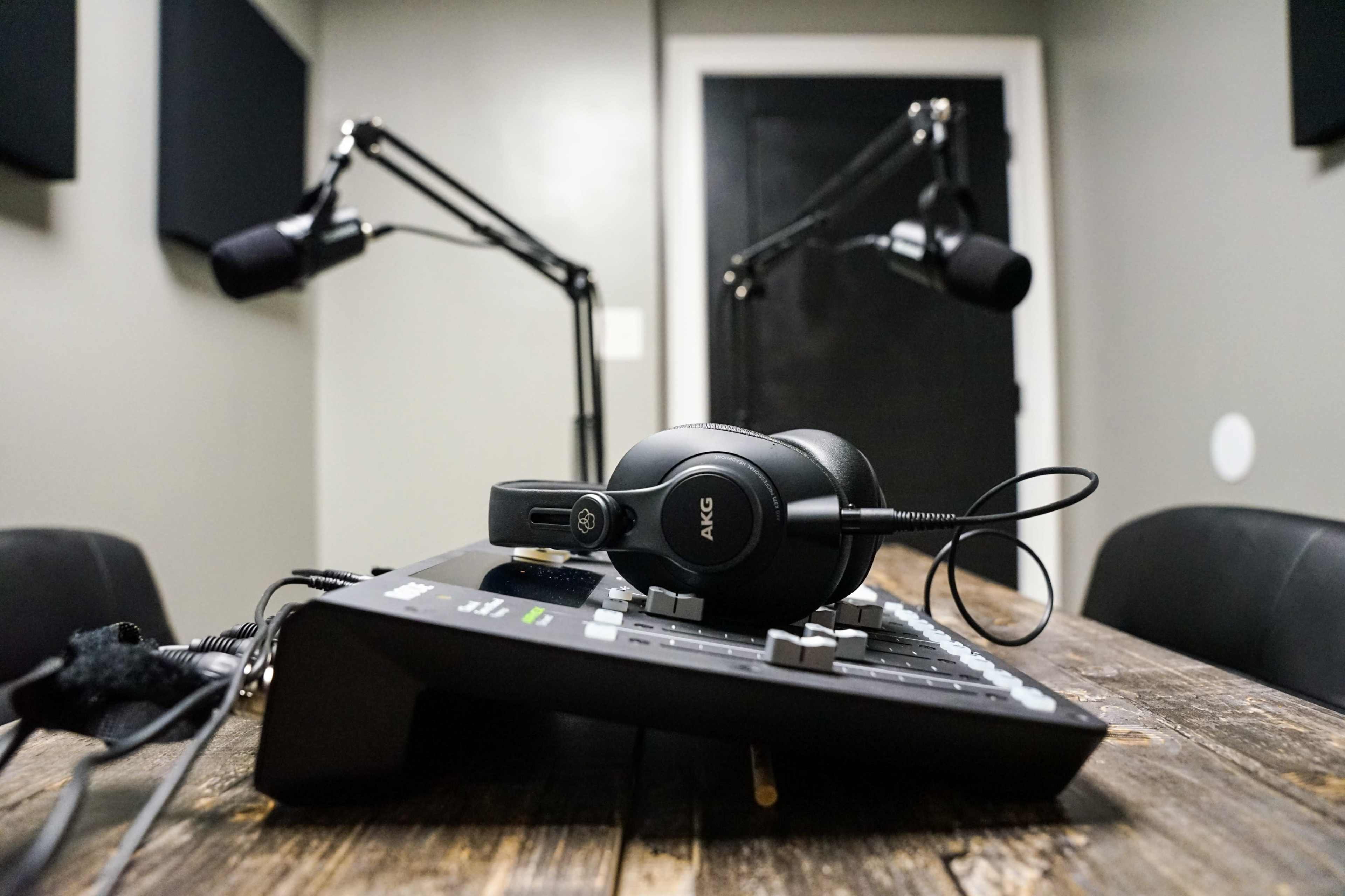 A black audio mixer with headphones sits on a wooden table in a sound recording studio featuring two microphones on adjustable arms.
