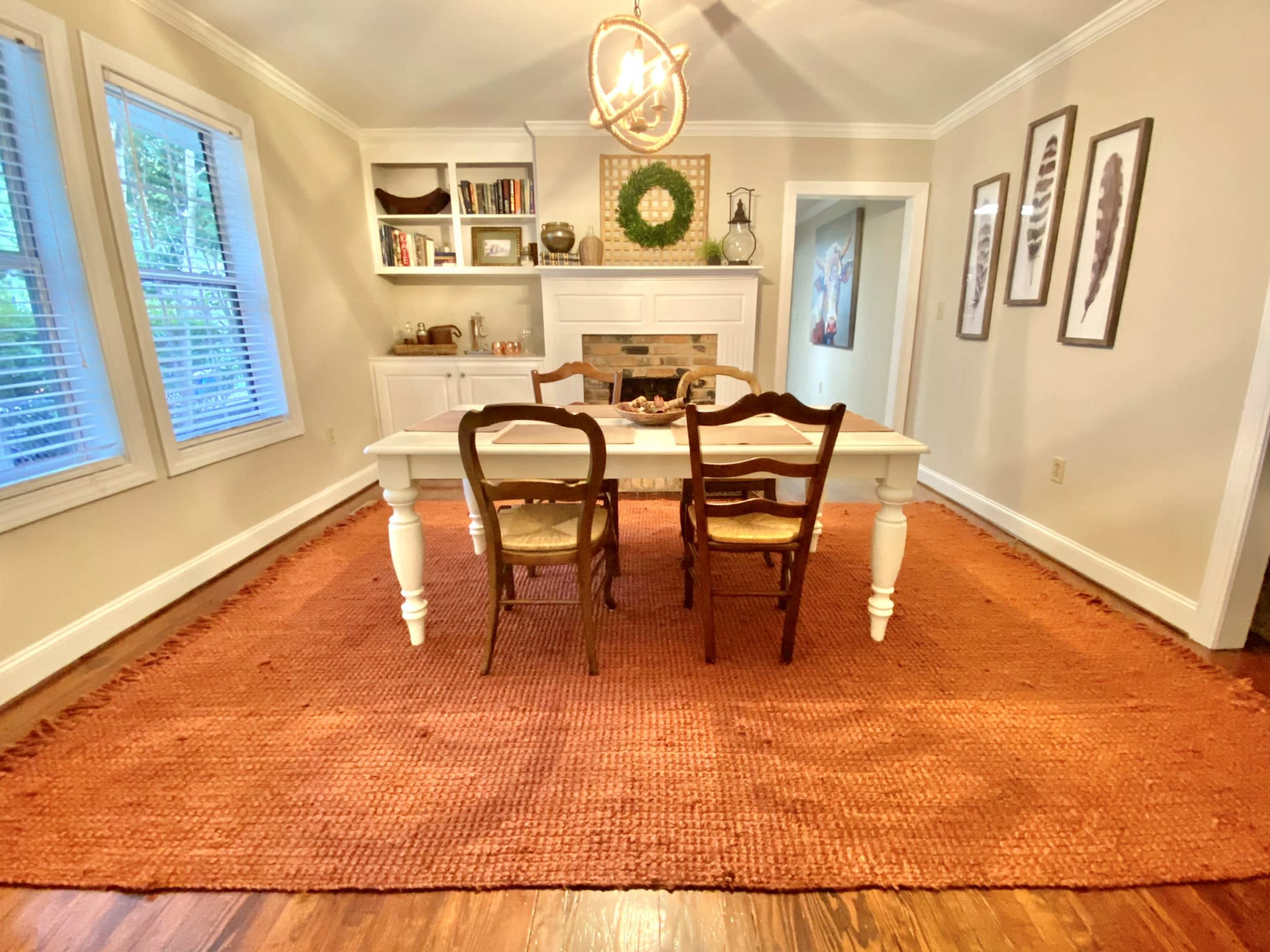 A dining room with a rectangular table surrounded by four wooden chairs, a large orange rug on the floor, and a fireplace with decorations above it.