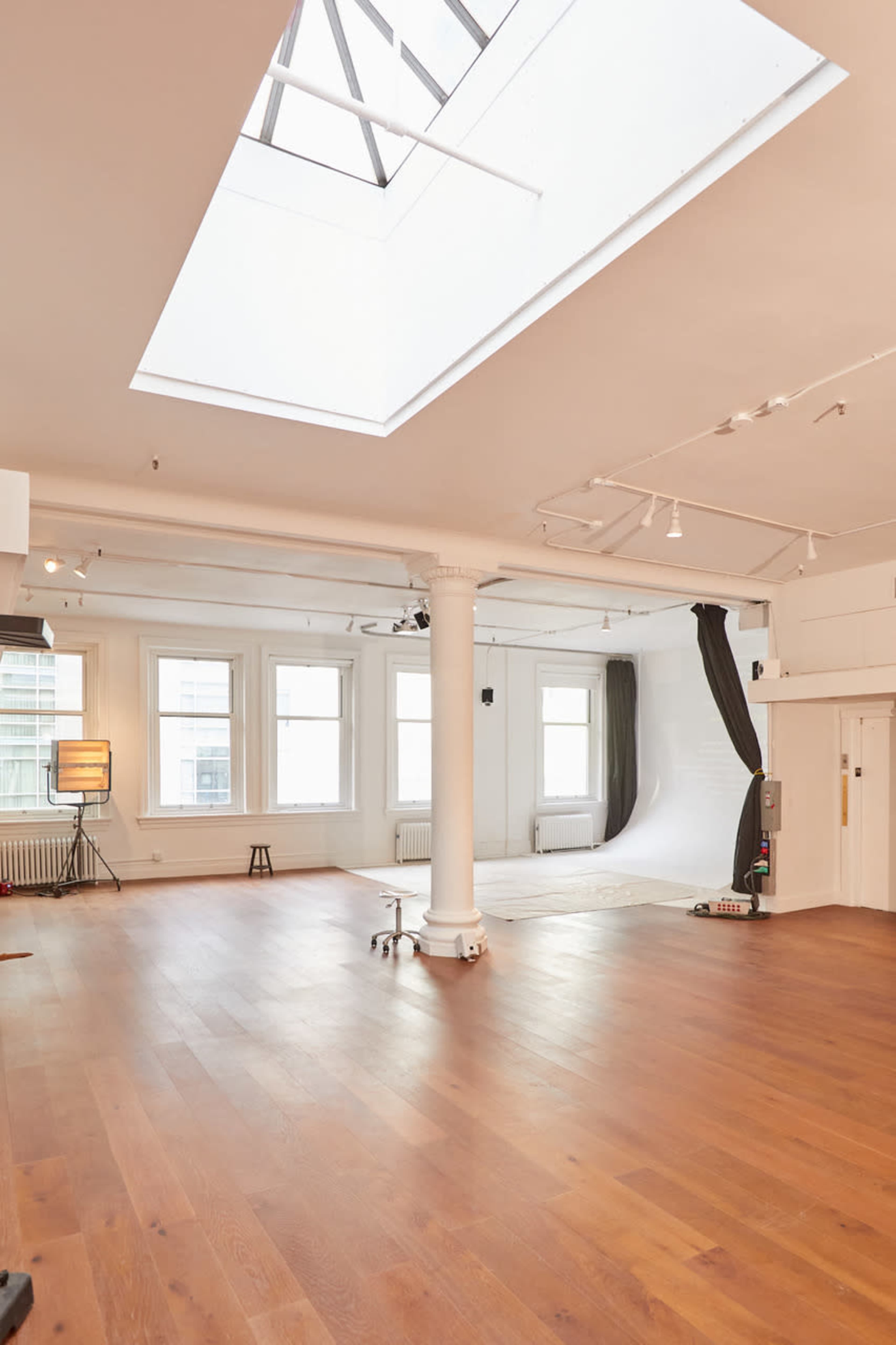 The image shows a spacious, well-lit studio with large windows, wooden flooring, and a backdrop area featuring a draped curtain.