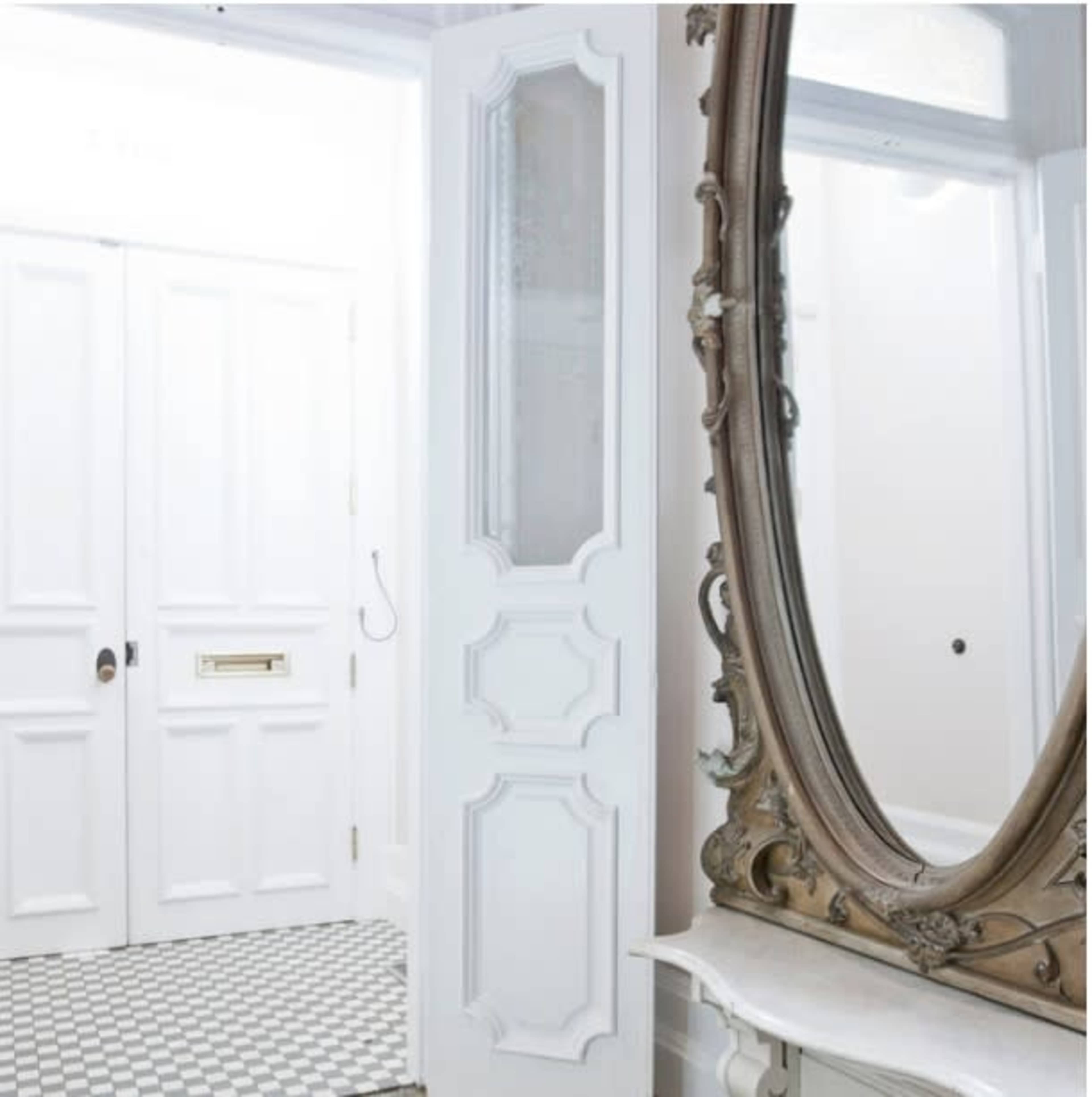 A bright hallway featuring a large ornate mirror and double white doors, with a patterned floor visible.
