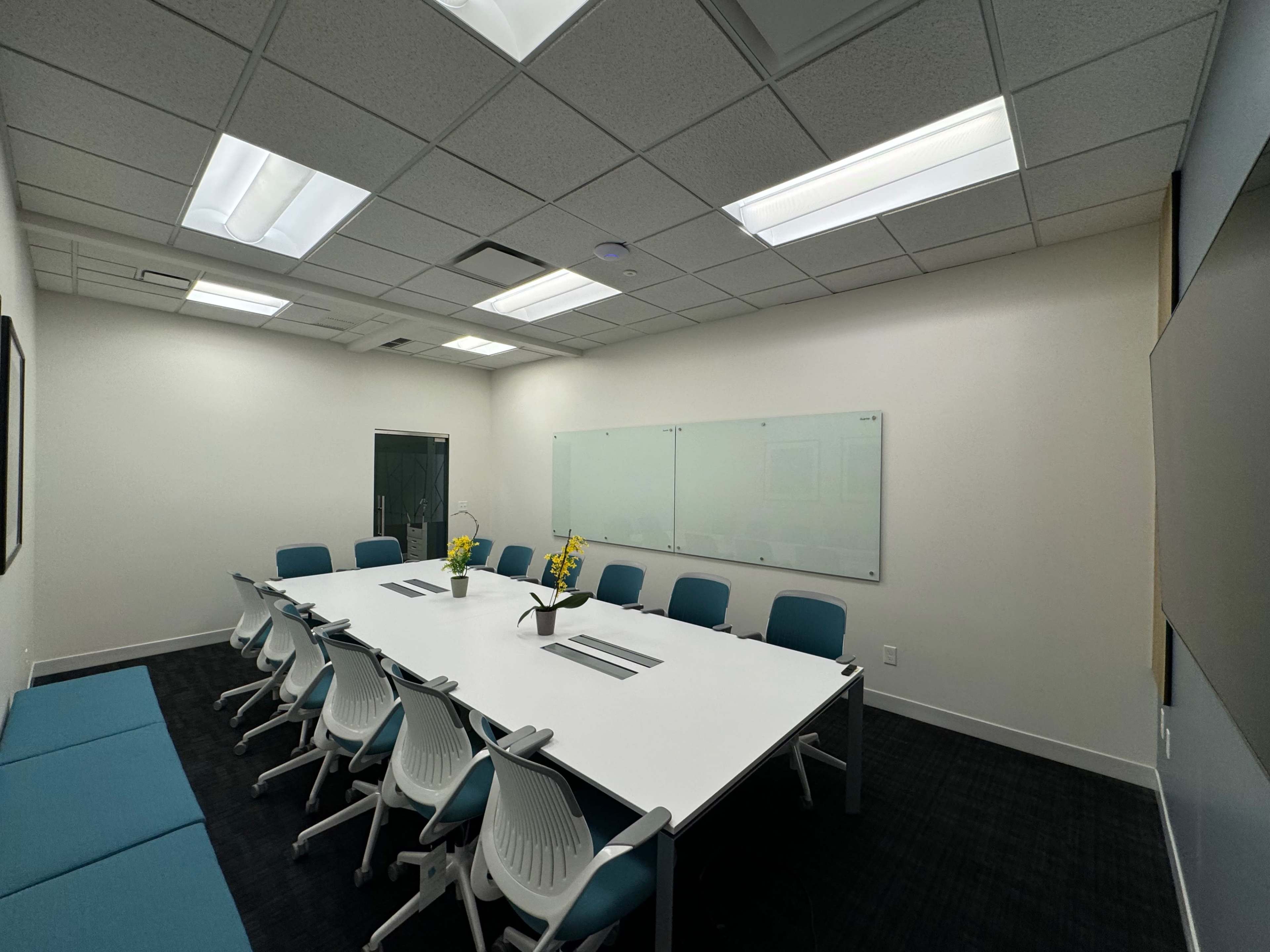 A conference room features a large white table surrounded by blue and white chairs, with a glass whiteboard on one wall and overhead lighting.