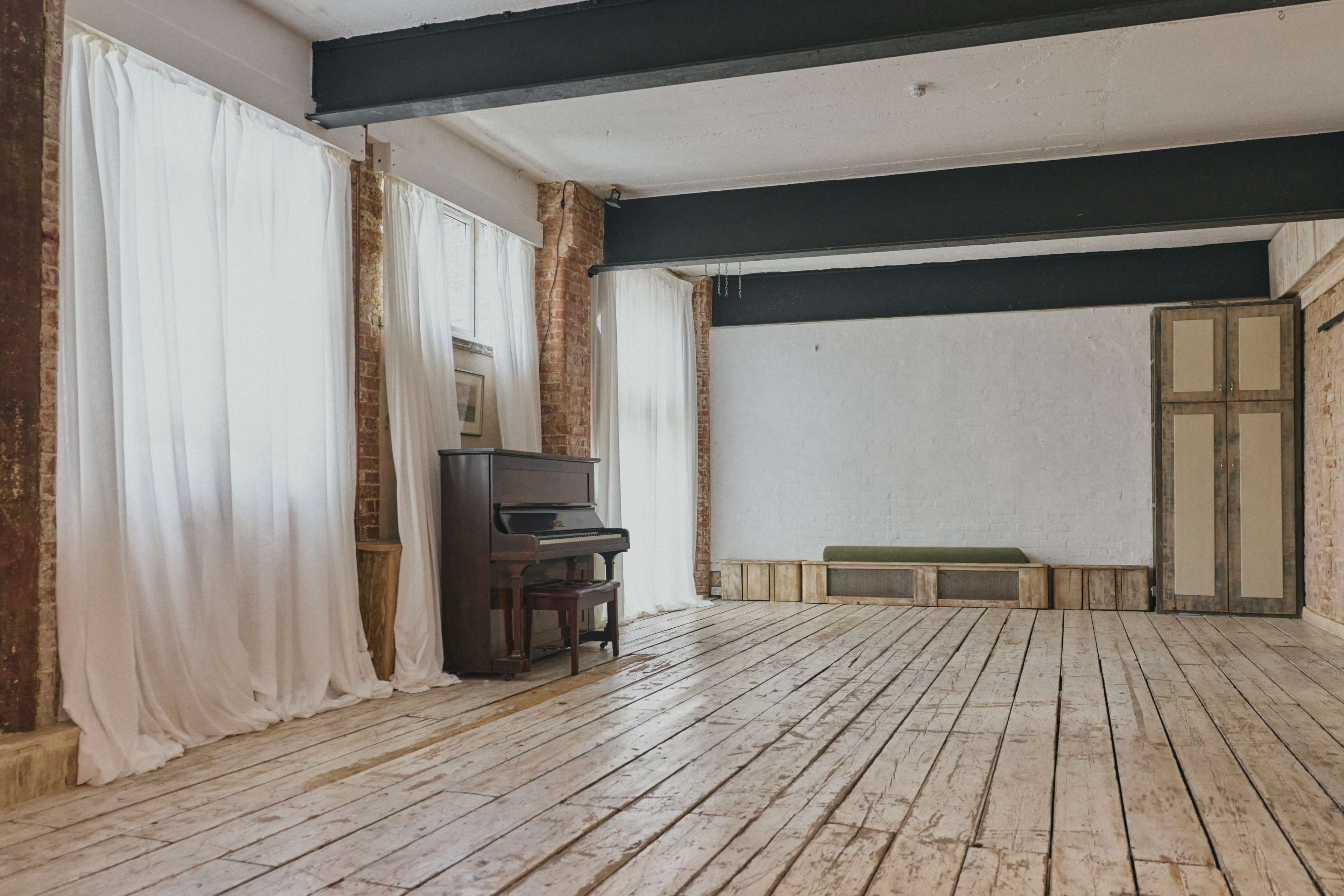 The image shows a spacious, minimalist room with wooden floors, a piano in the corner, large windows covered with sheer curtains, and exposed brick walls.