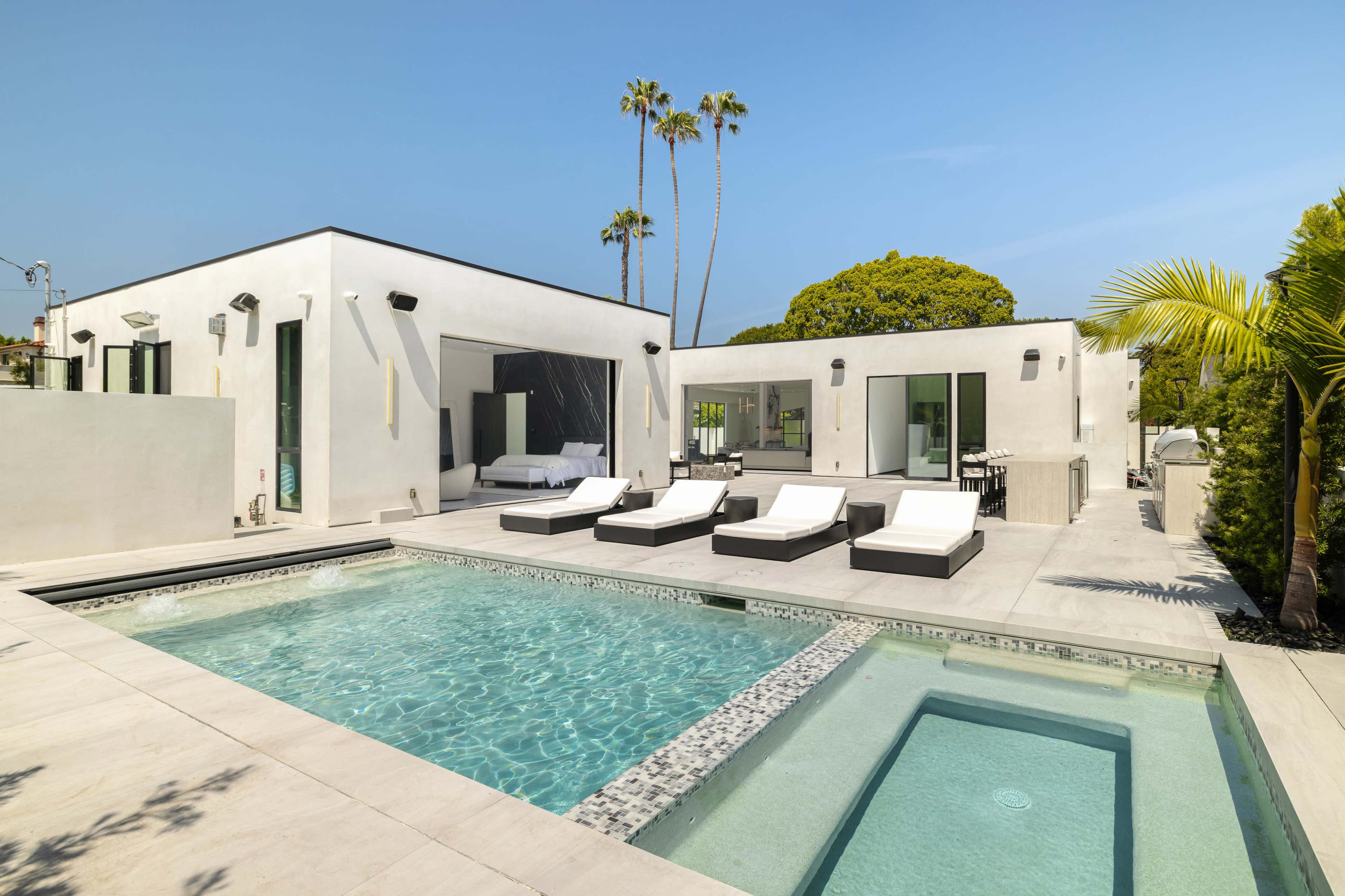 The image shows a modern home with a swimming pool, lounge chairs, and palm trees under a clear blue sky.