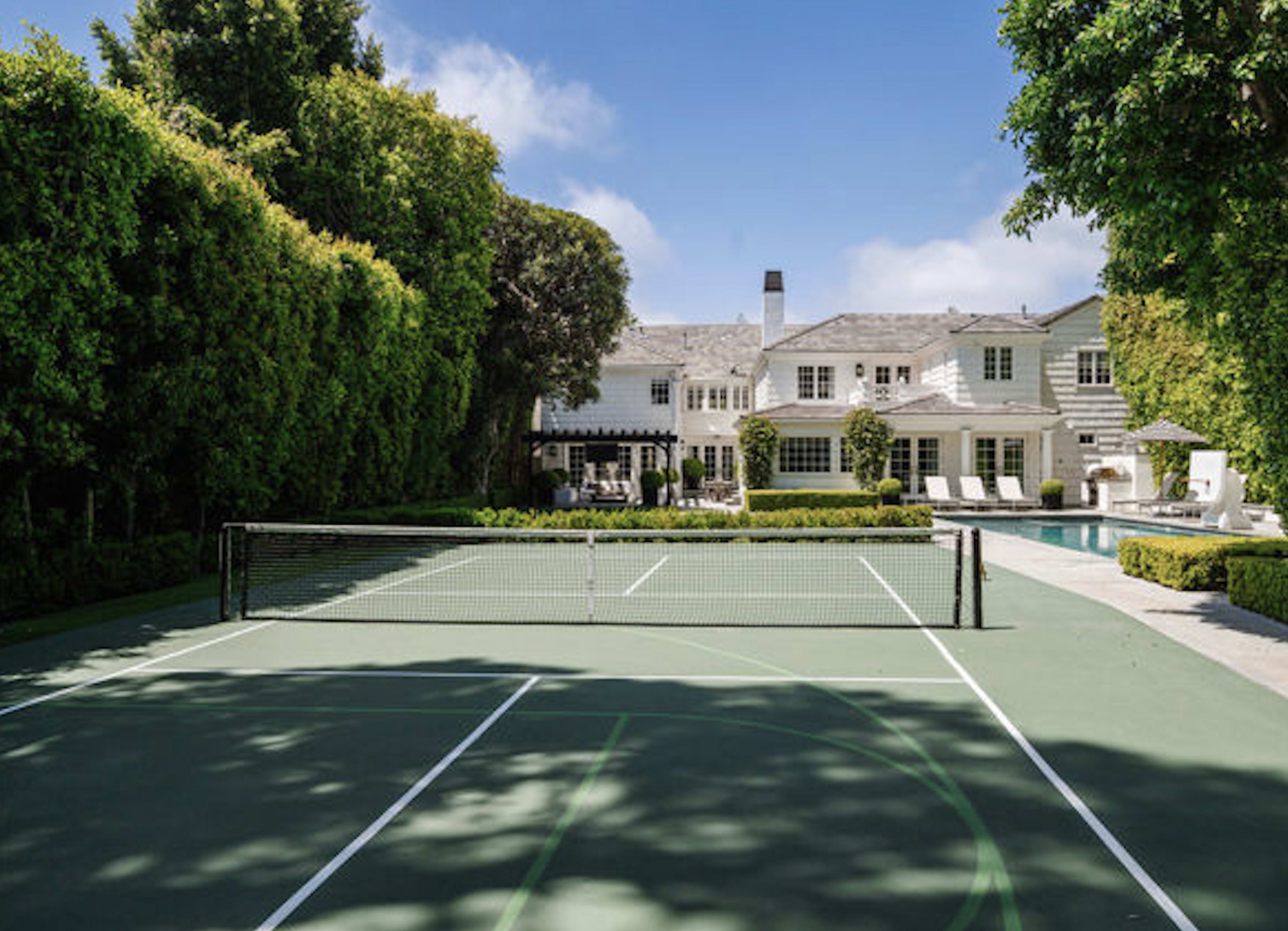 The image shows a tennis court surrounded by lush greenery, with a large house and swimming pool visible in the background.