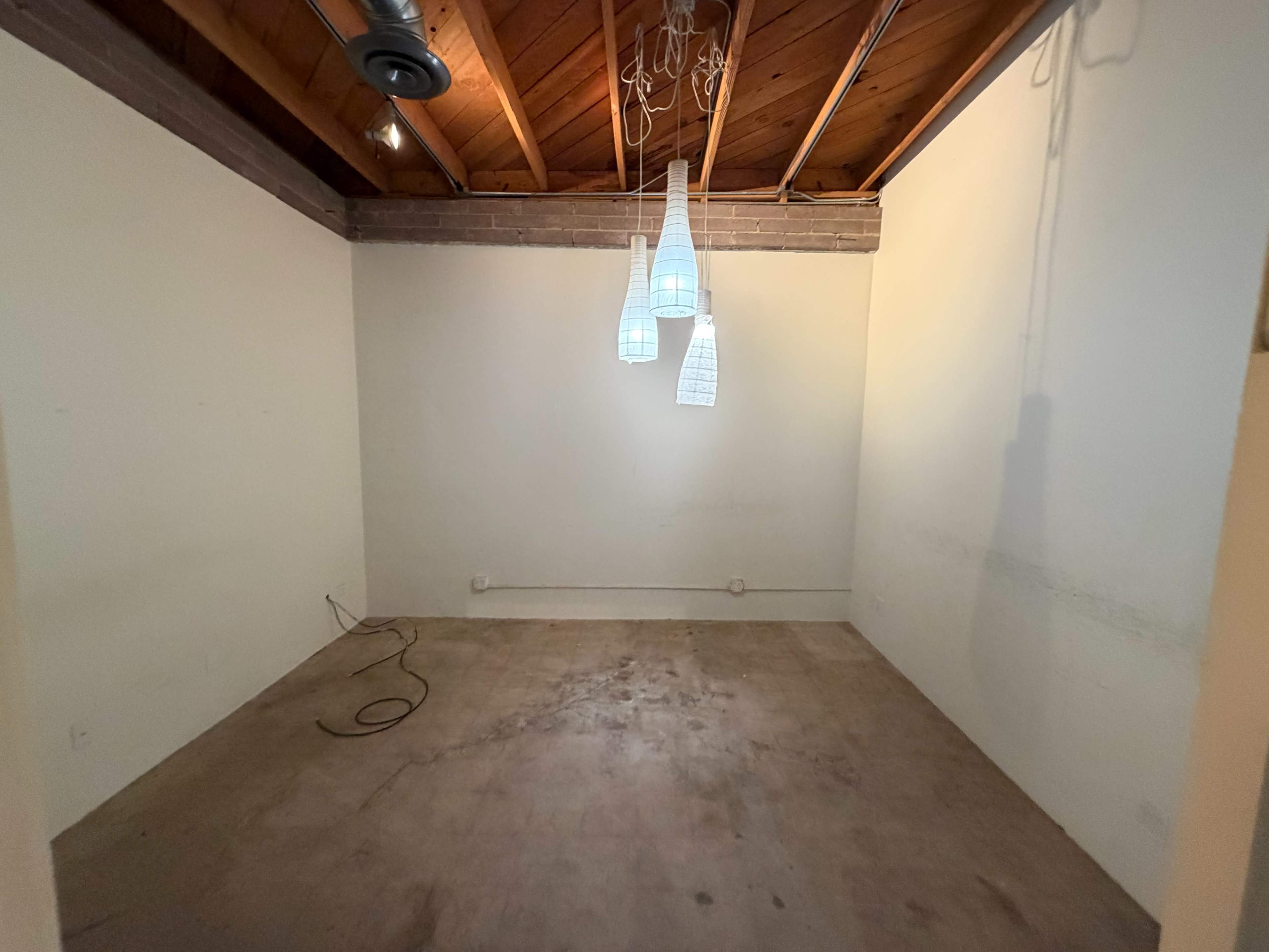 The image shows a sparsely furnished room with bare walls, a concrete floor, and three hanging light fixtures from the ceiling.