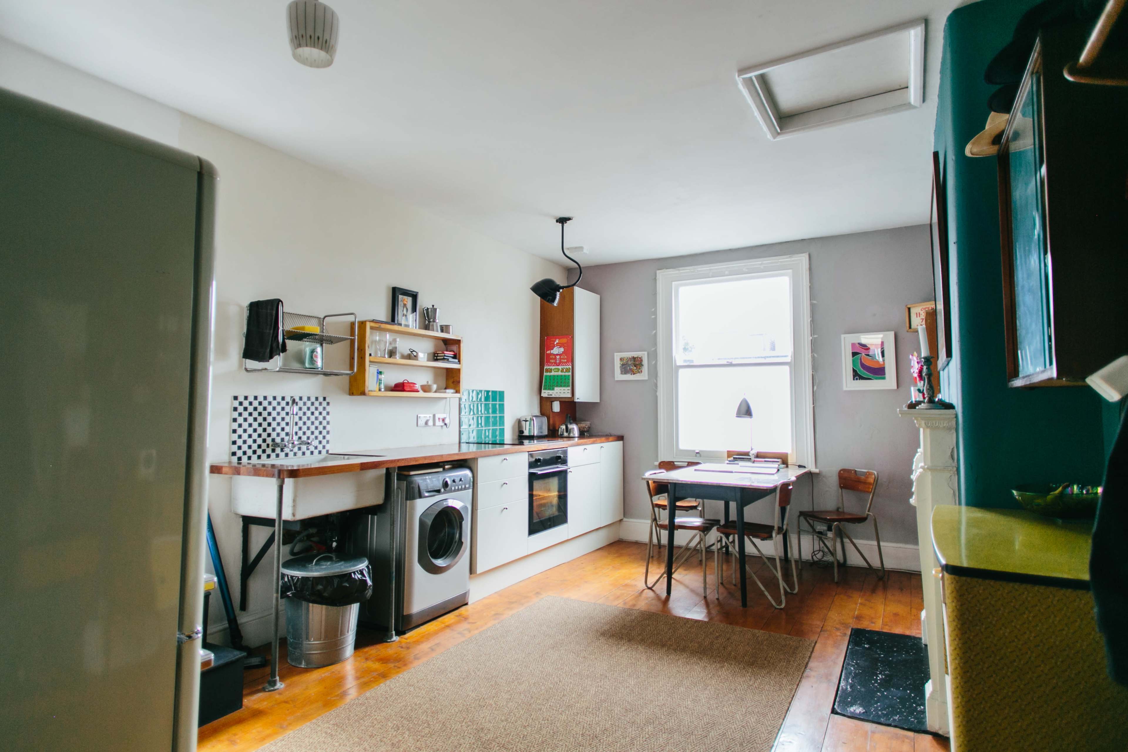 A kitchen features a washing machine, wooden shelves with kitchen items, a dining table with chairs, and a window allowing natural light.