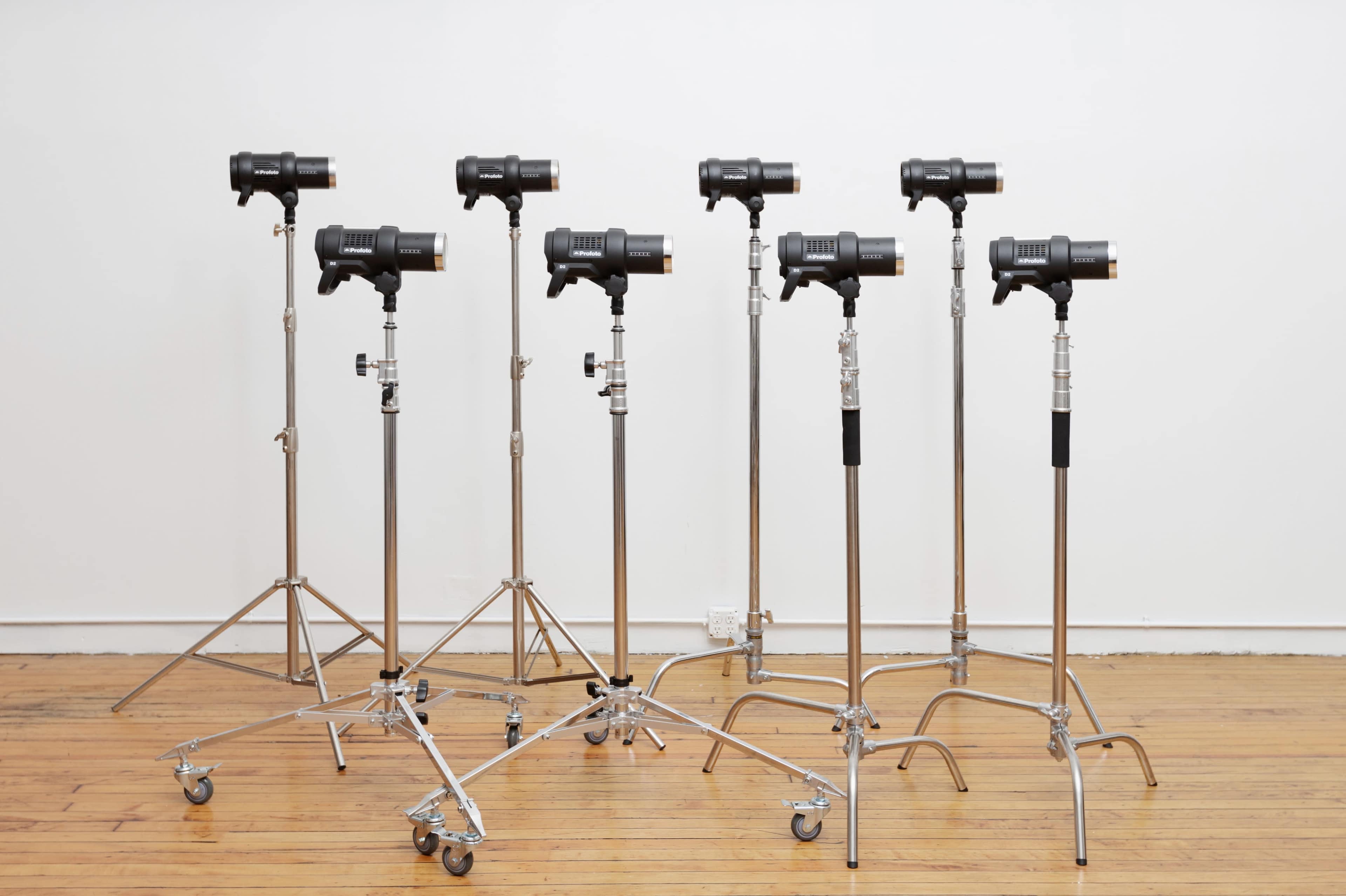 A row of eight studio lights mounted on adjustable stands is set against a plain white wall on a wooden floor.