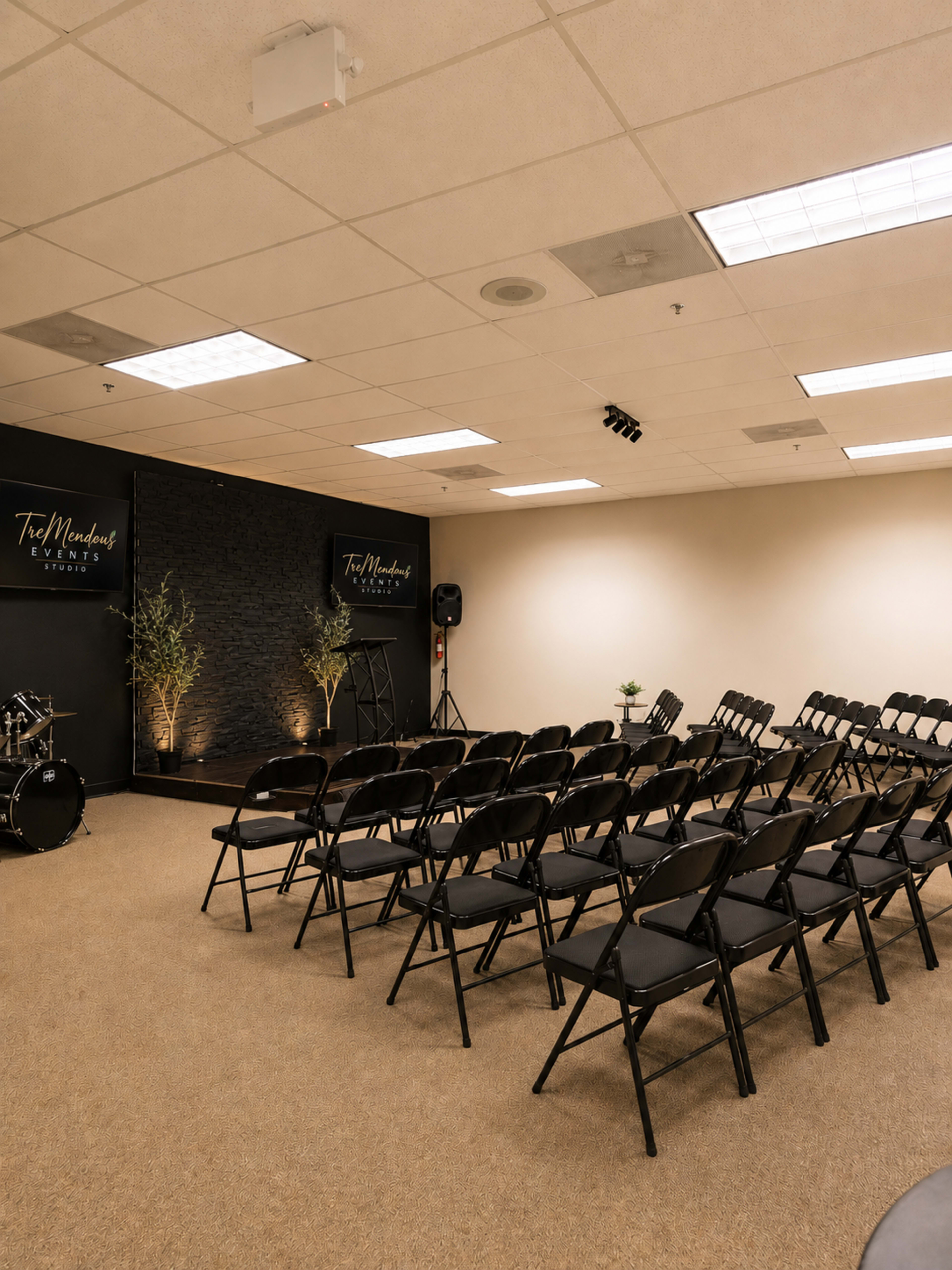 The image shows a spacious room set up with rows of black folding chairs facing a dark wall featuring a logo, with a small drum set and plants in the corners.