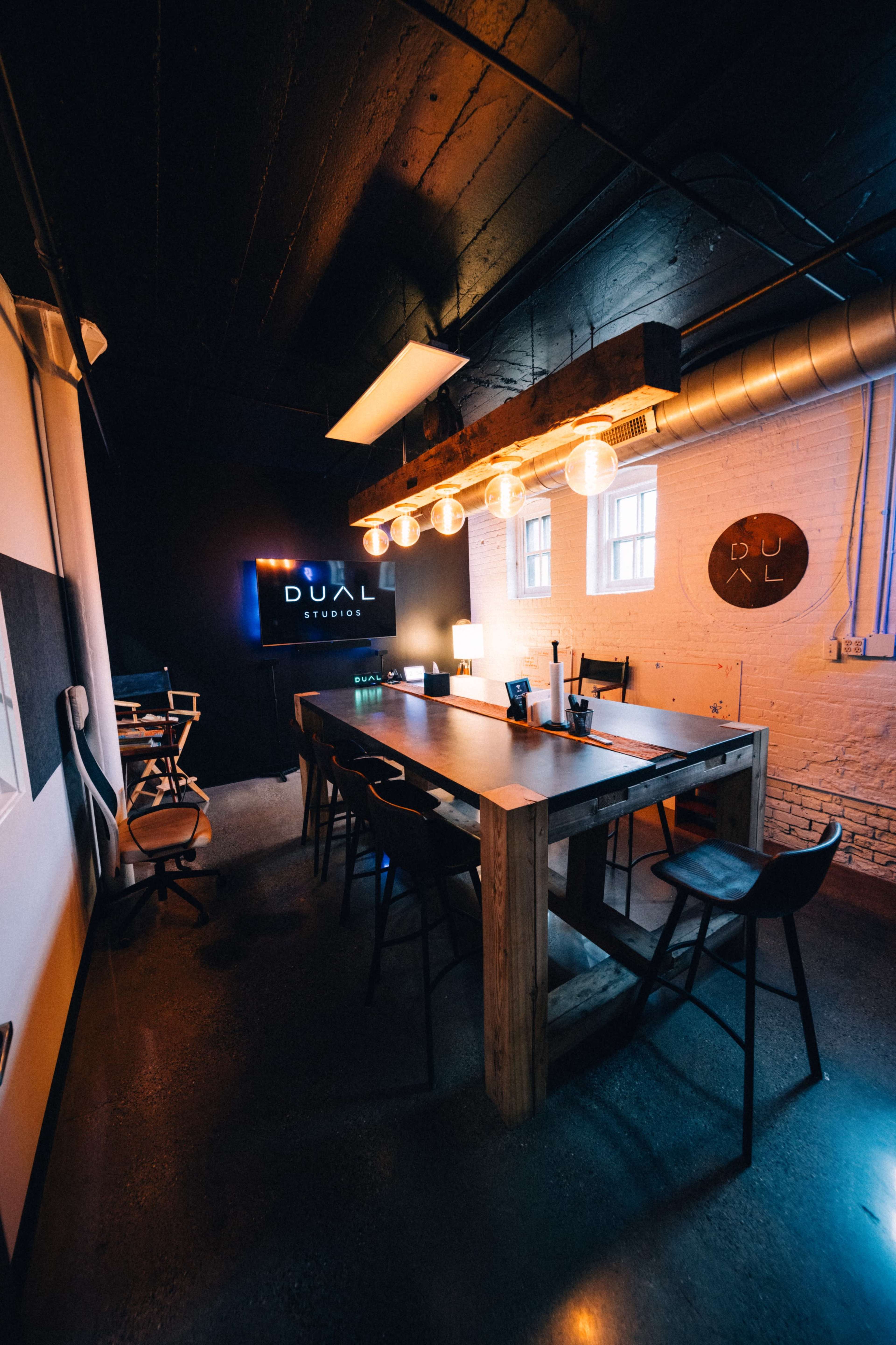 A modern conference room features a long wooden table surrounded by chairs, with a dark wall displaying the "DUAL" studio logo and decorative lighting.