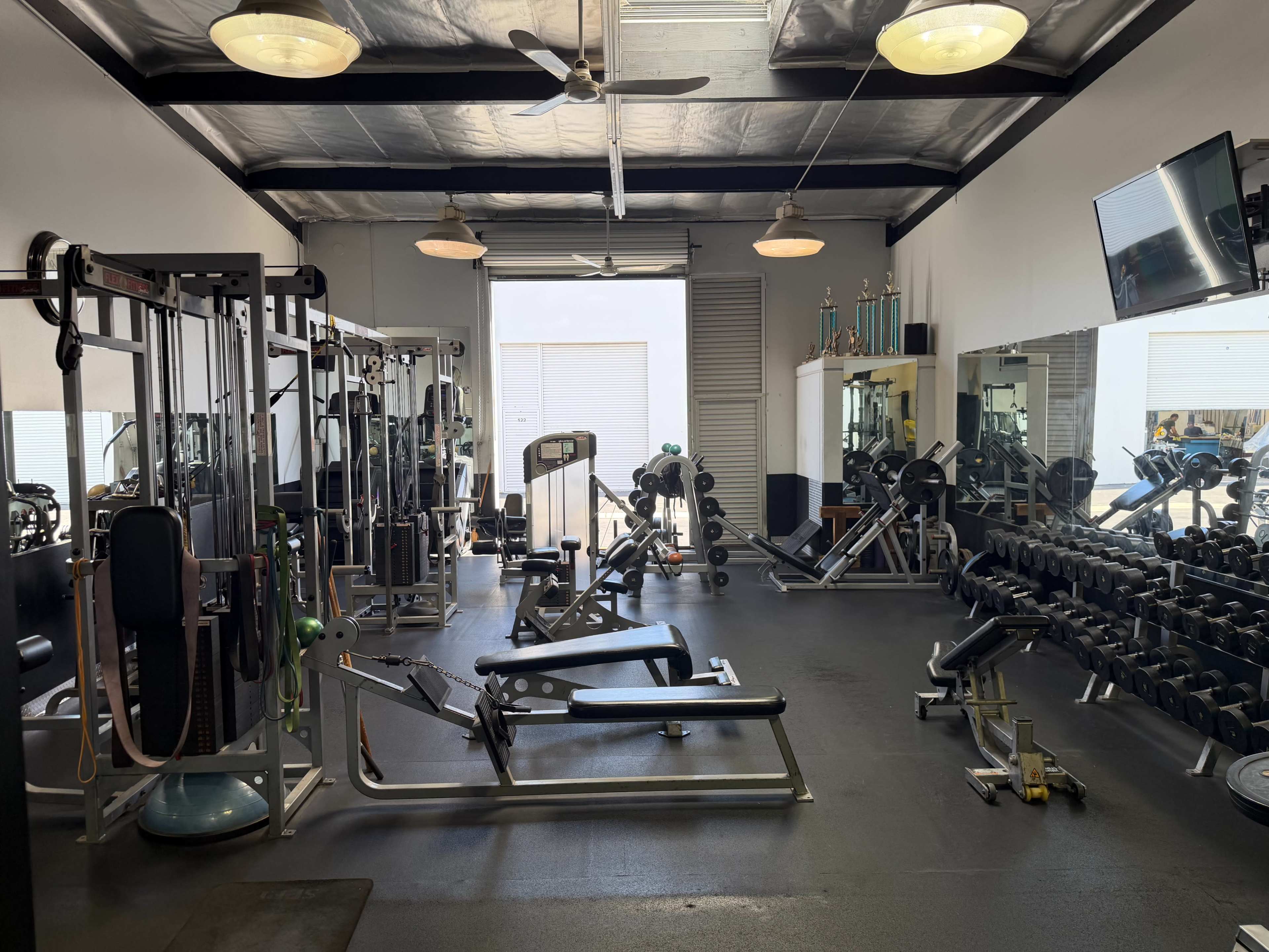 The image shows a well-equipped gym featuring various exercise machines, free weights, mirrors, and an open entrance.