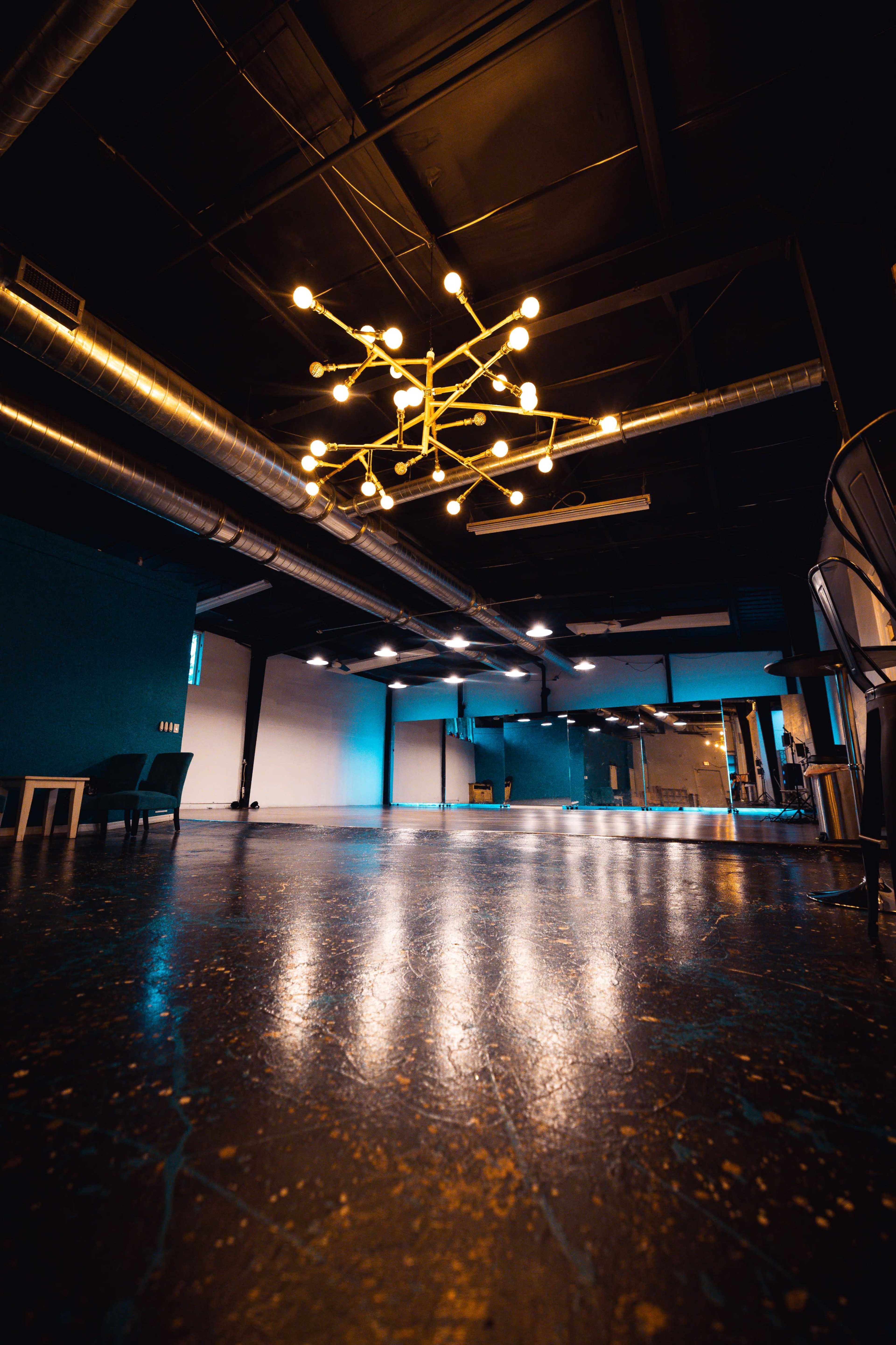 The image shows a spacious, empty room with a polished floor, industrial lighting, and a large chandelier hanging from the ceiling.