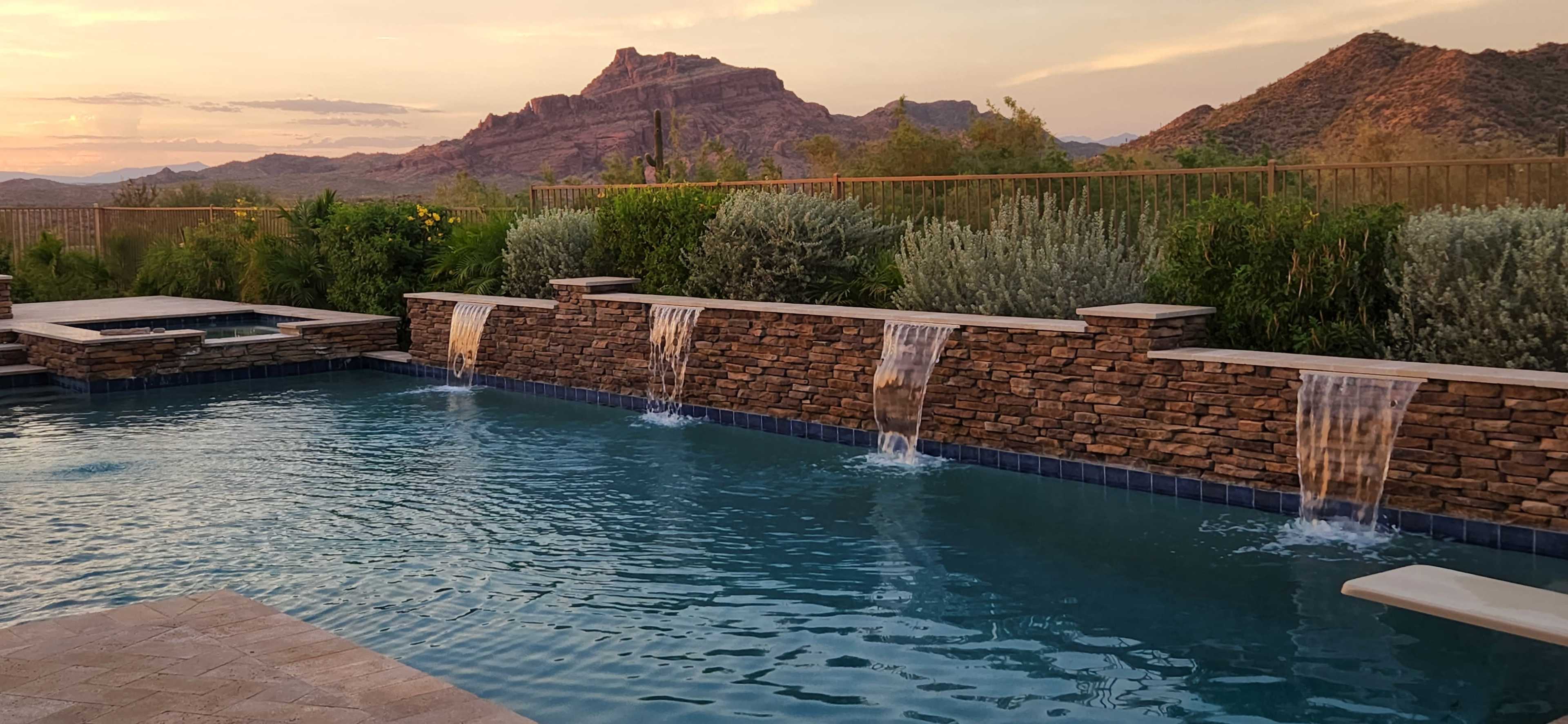 A swimming pool with stone waterfalls is situated in a landscaped area, framed by mountains in the background during sunset.