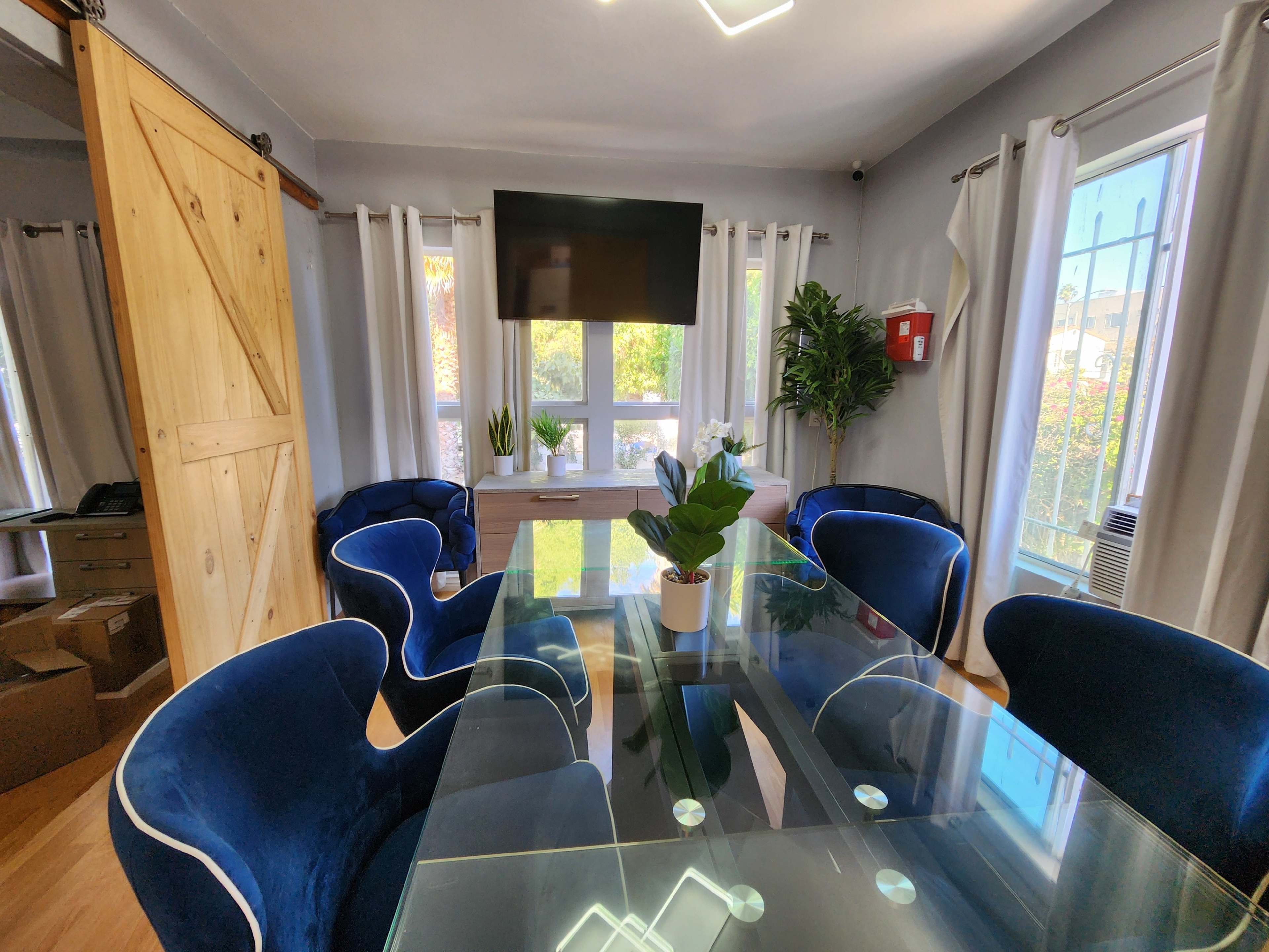 The image shows a modern dining area with a glass table surrounded by blue velvet chairs, a large television mounted on the wall, and potted plants near the windows.