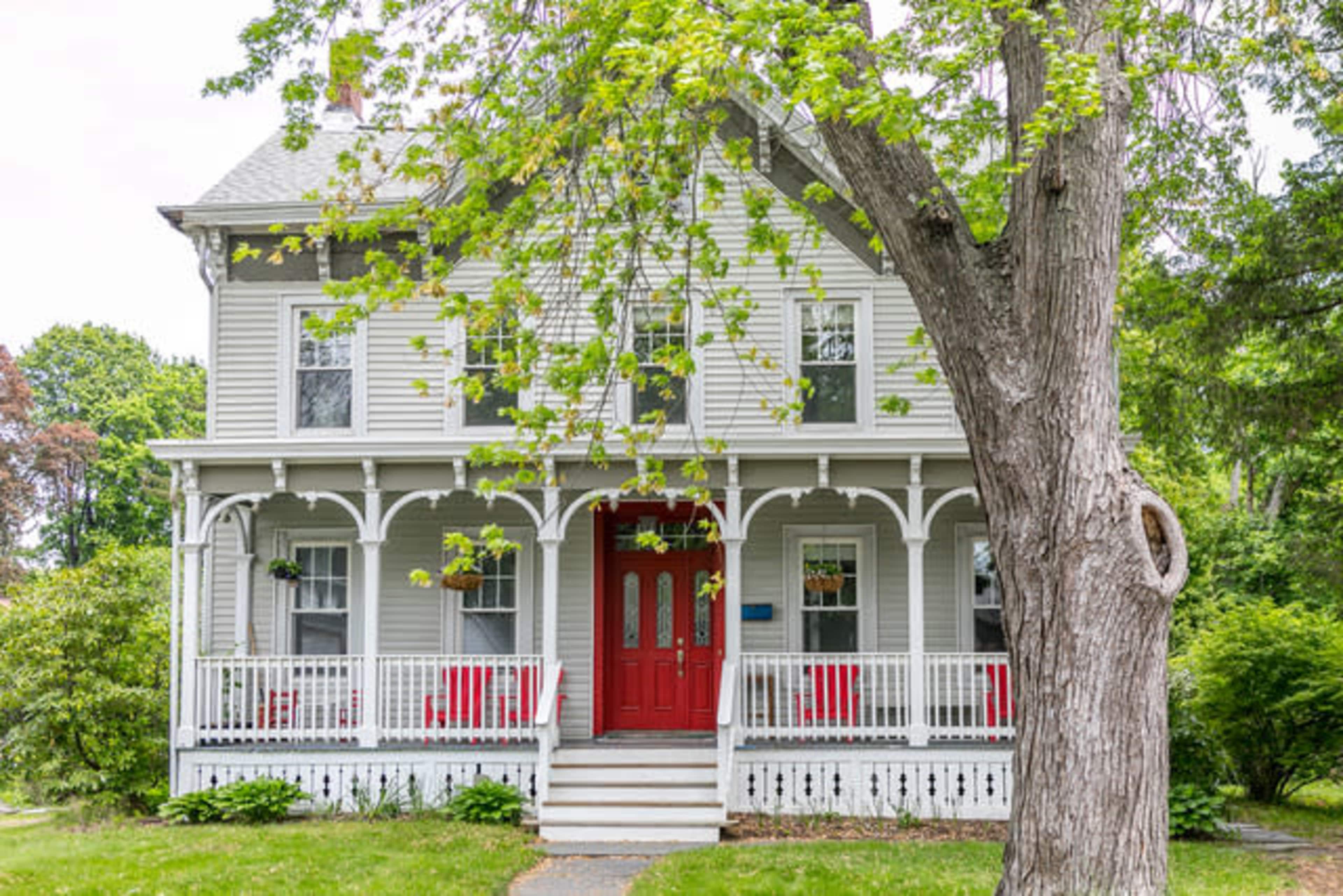 A two-story house with a gray exterior, white trim, and a bright red front door is surrounded by a tree and lush greenery.