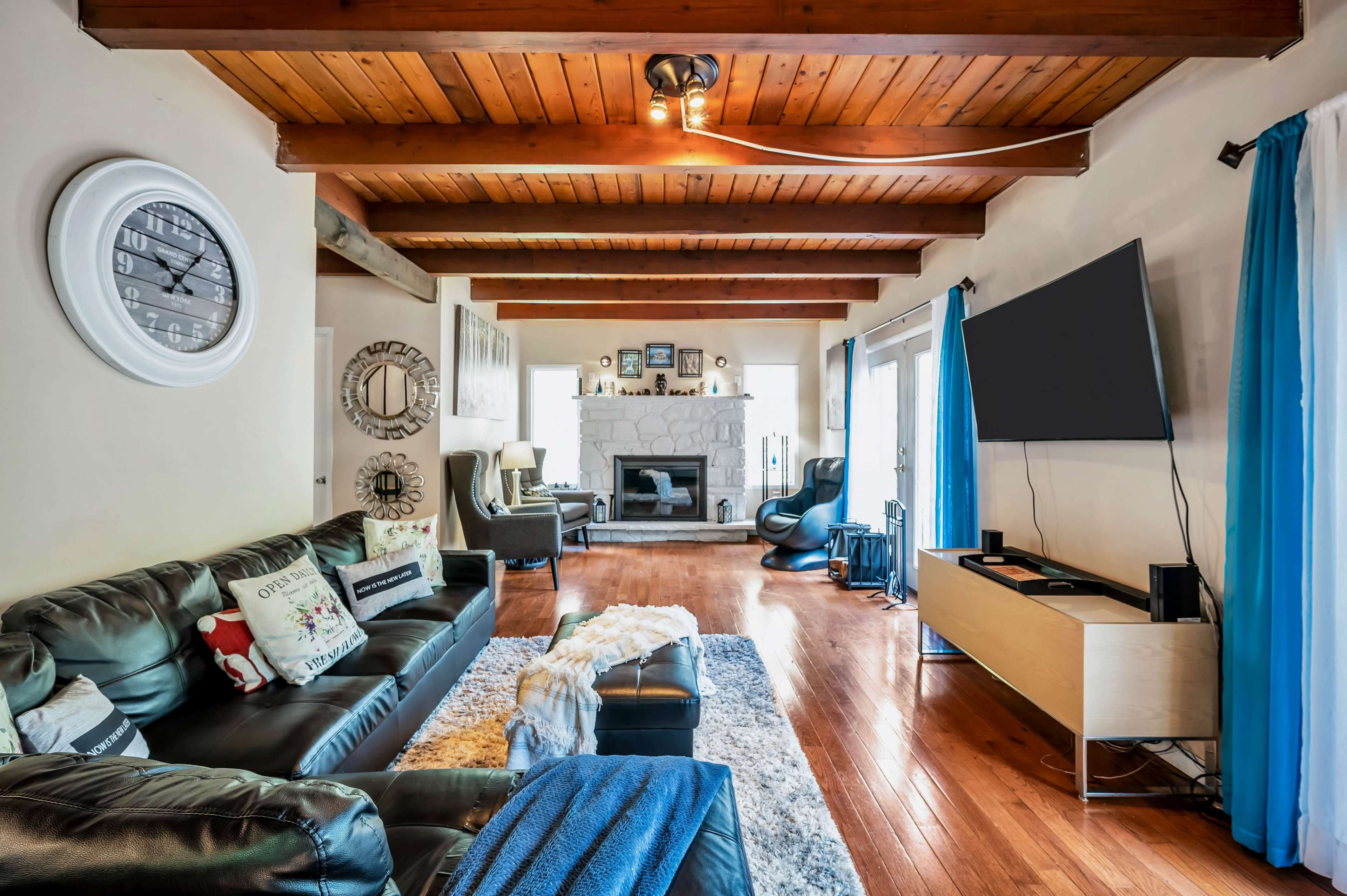 The image shows a modern living room featuring leather sofas, a wooden ceiling, a stone fireplace, and a large wall-mounted television.