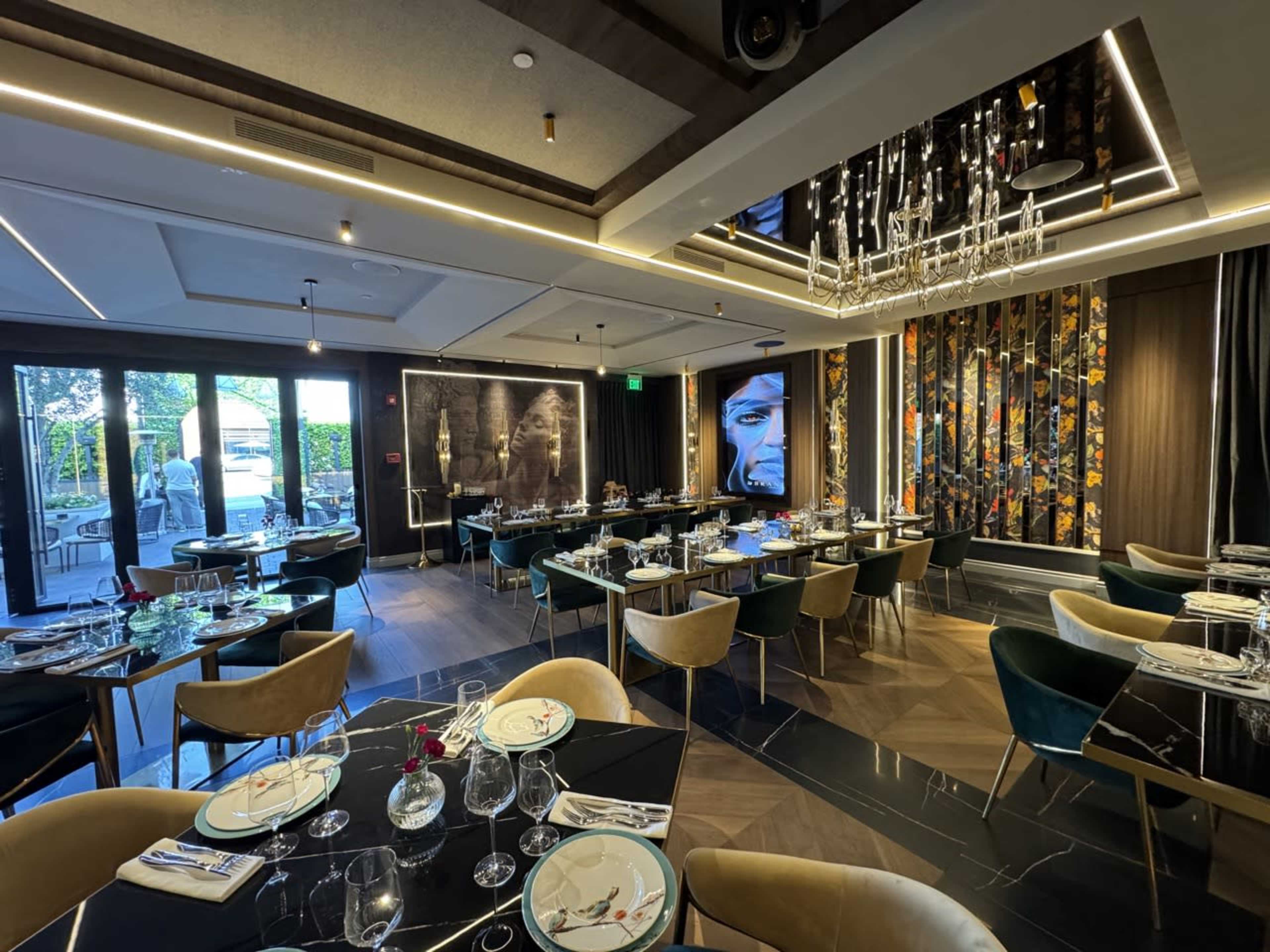 The image shows a modern dining room with elegant seating, a chandelier, and large windows that allow natural light to enter.