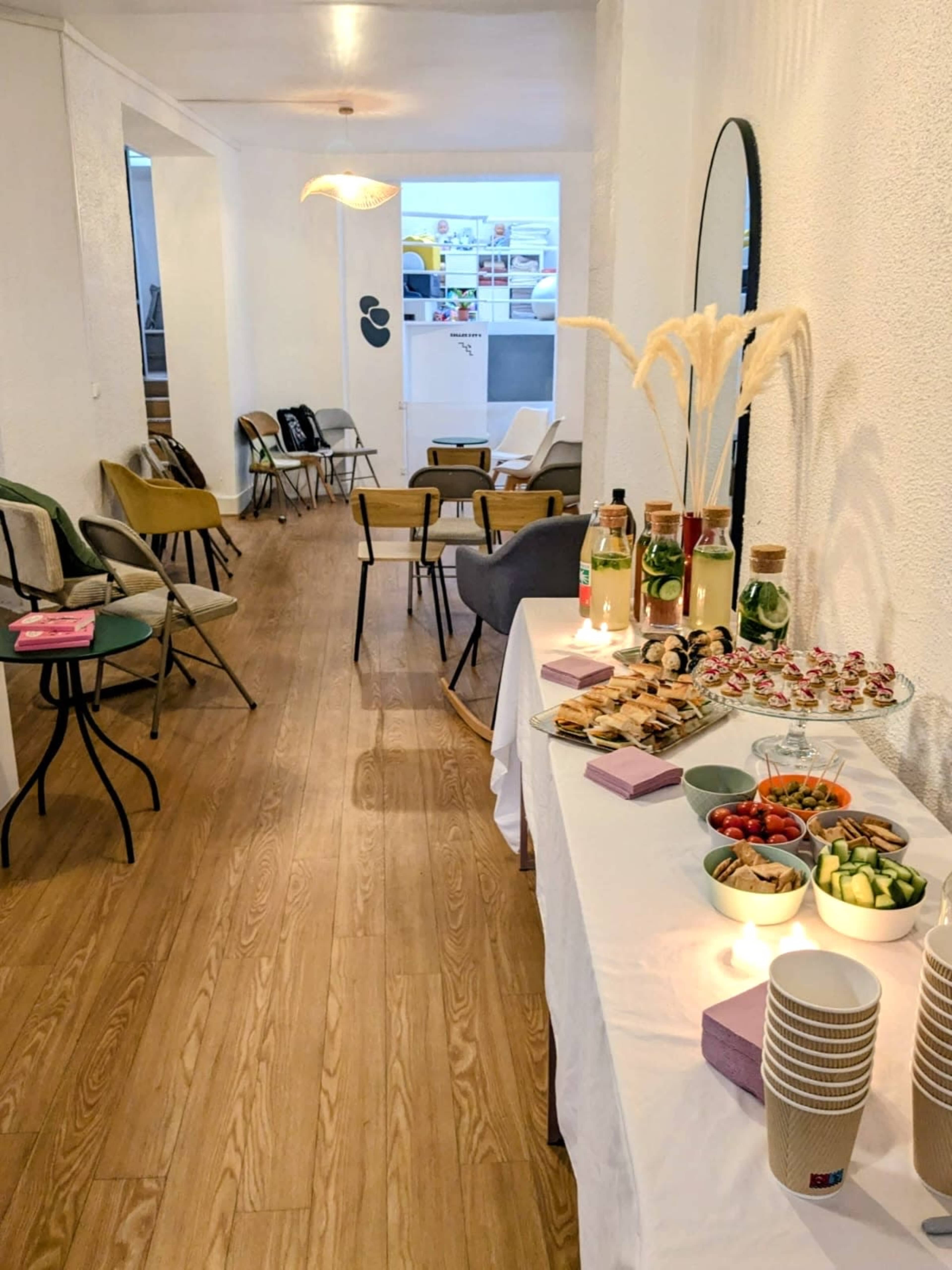 The image shows a spacious room with a long table set with various food items and drinks, surrounded by a mix of chairs arranged along the walls.