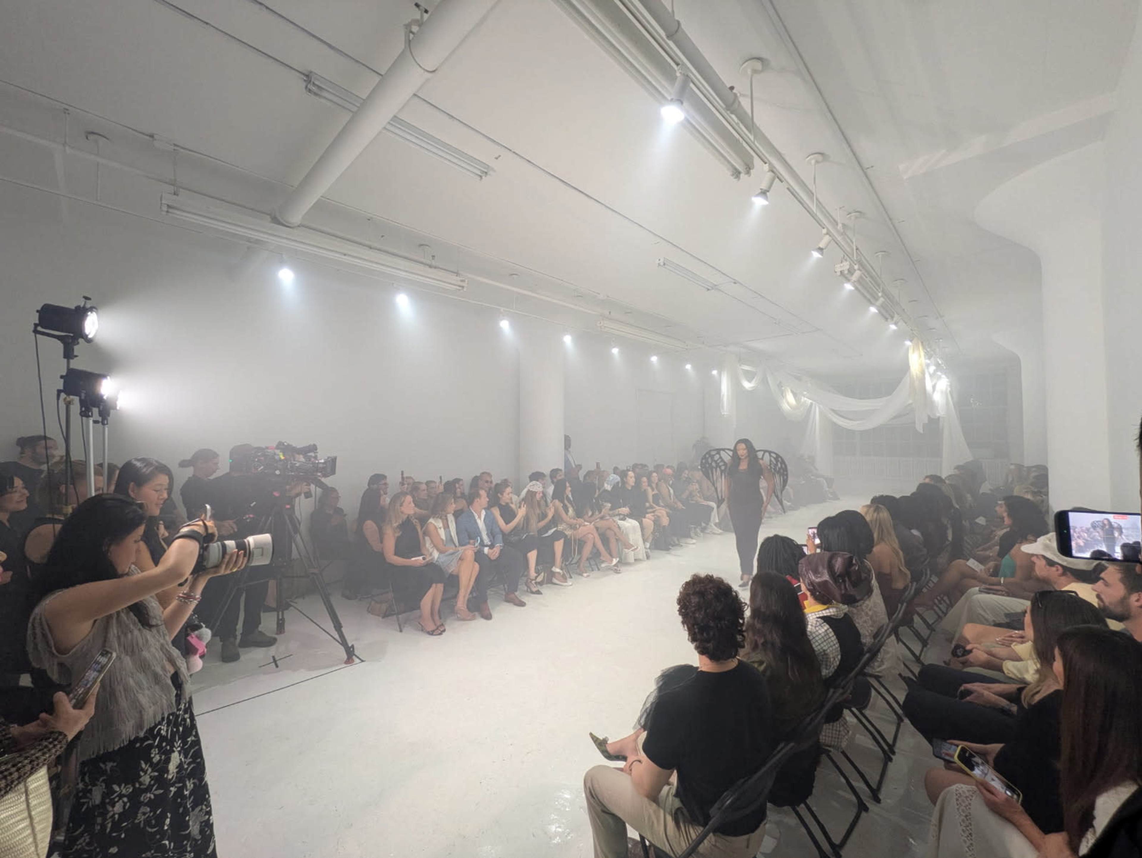 A fashion show takes place in a minimalist white studio, with a runway set up and an audience seated on either side, while photographers capture the event.