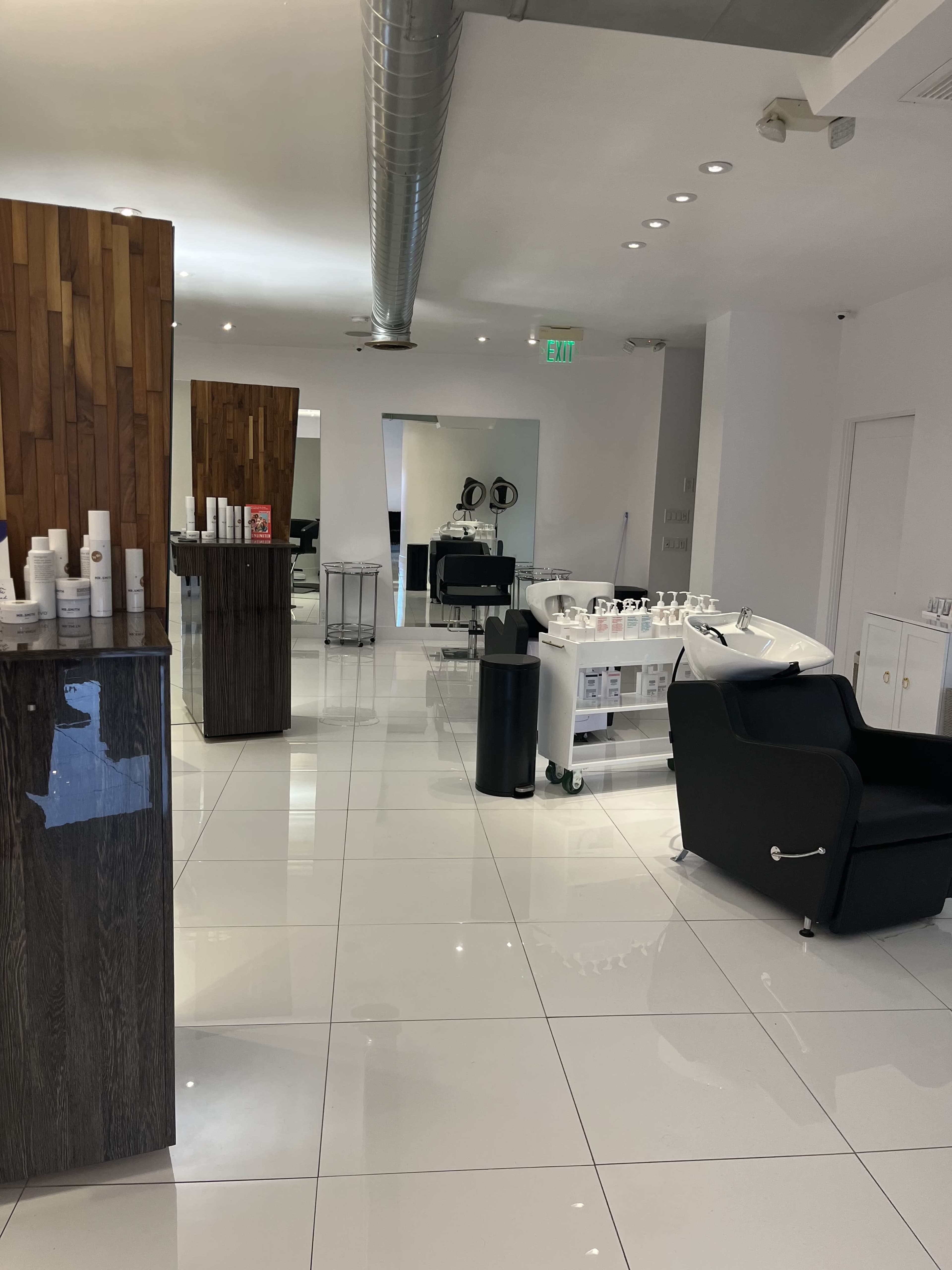A modern salon interior featuring sleek white tiles, a wooden accent wall, a black seating area, and a styling station with mirrors and salon equipment.