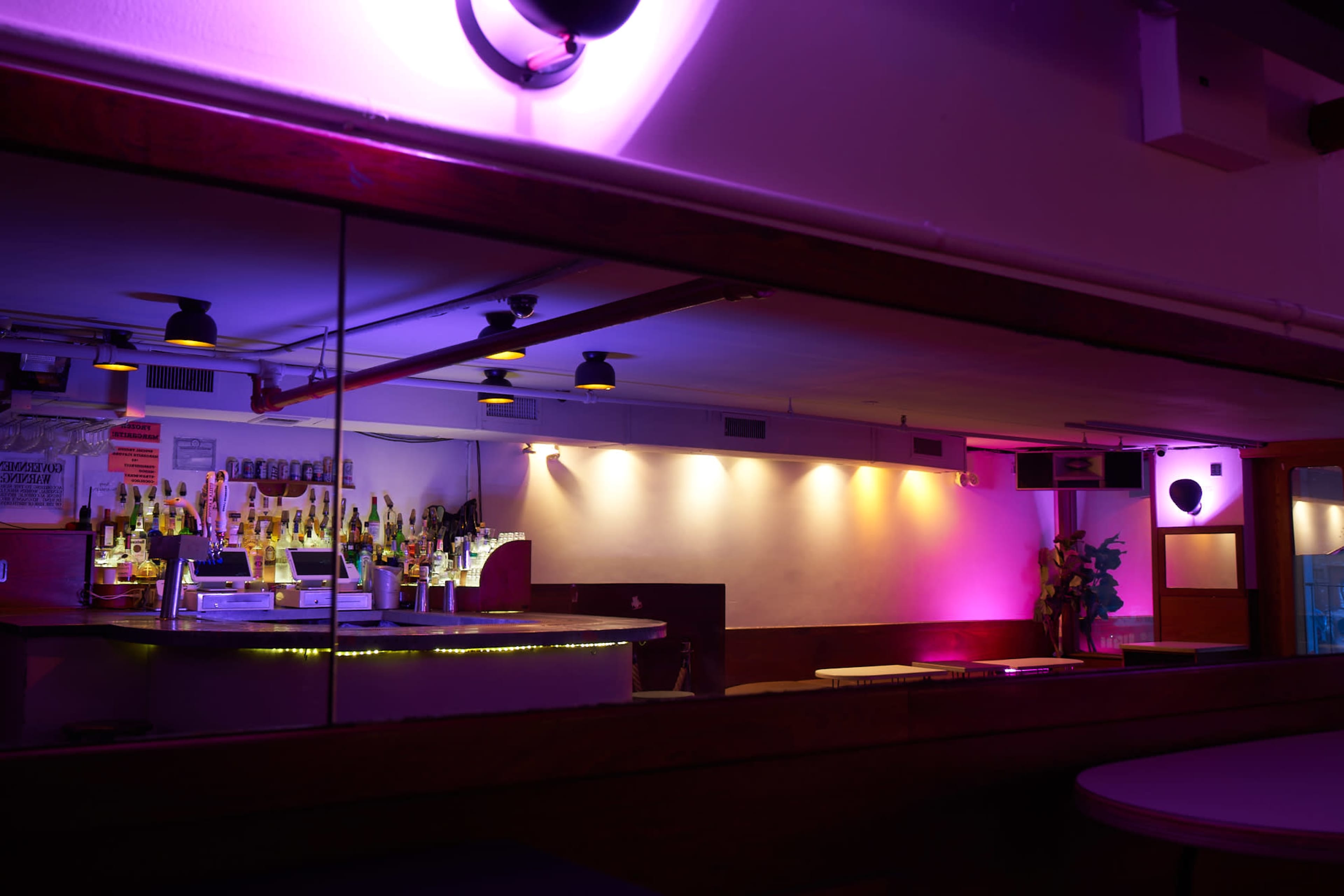 The image shows a dimly lit bar area with a mirrored wall and colorful lighting highlighting the shelves of bottles behind the counter.