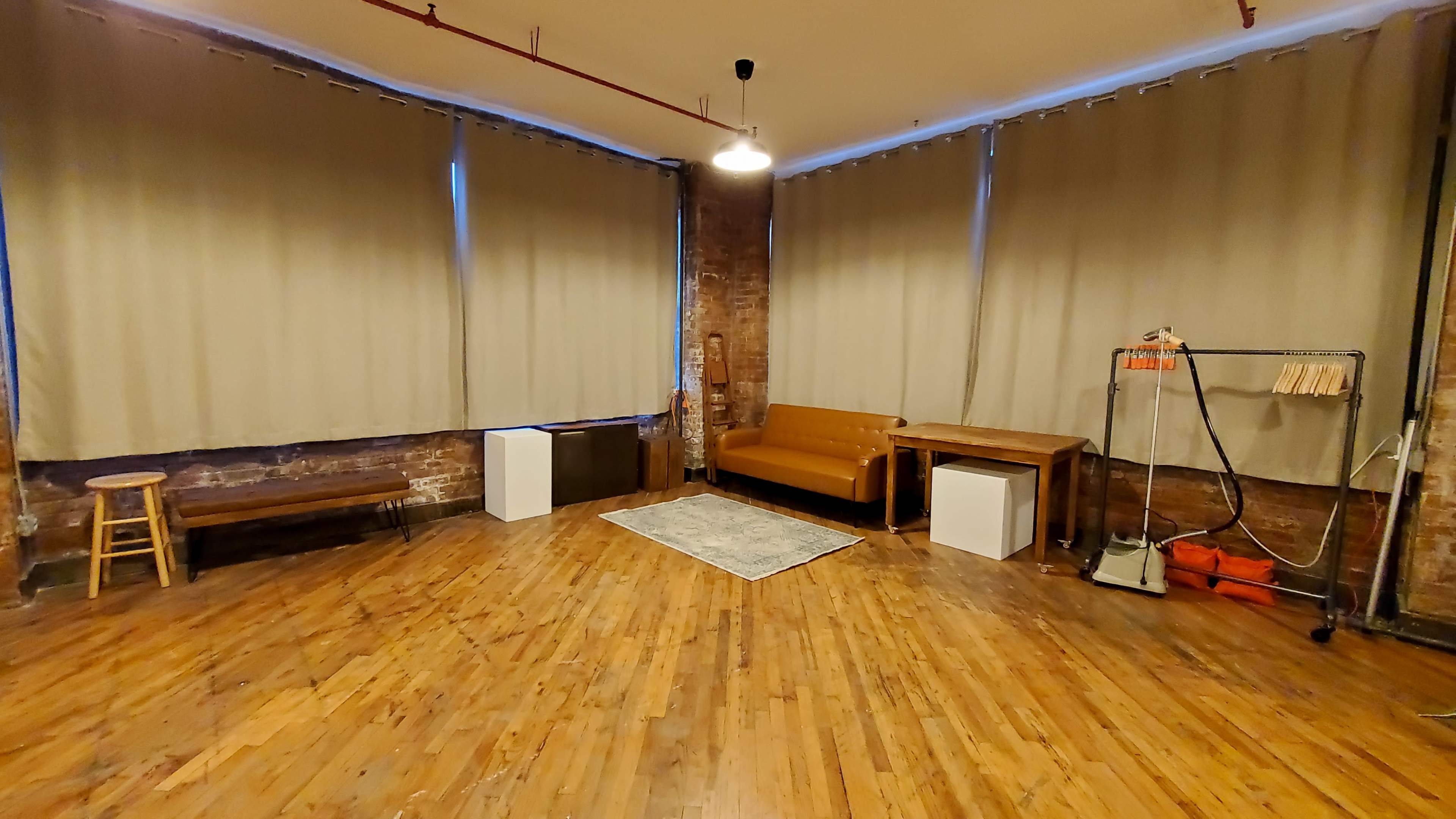 A spacious room with hardwood floors, large windows covered by beige curtains, a brown sofa, a wooden table, a stool, and a white cabinet.