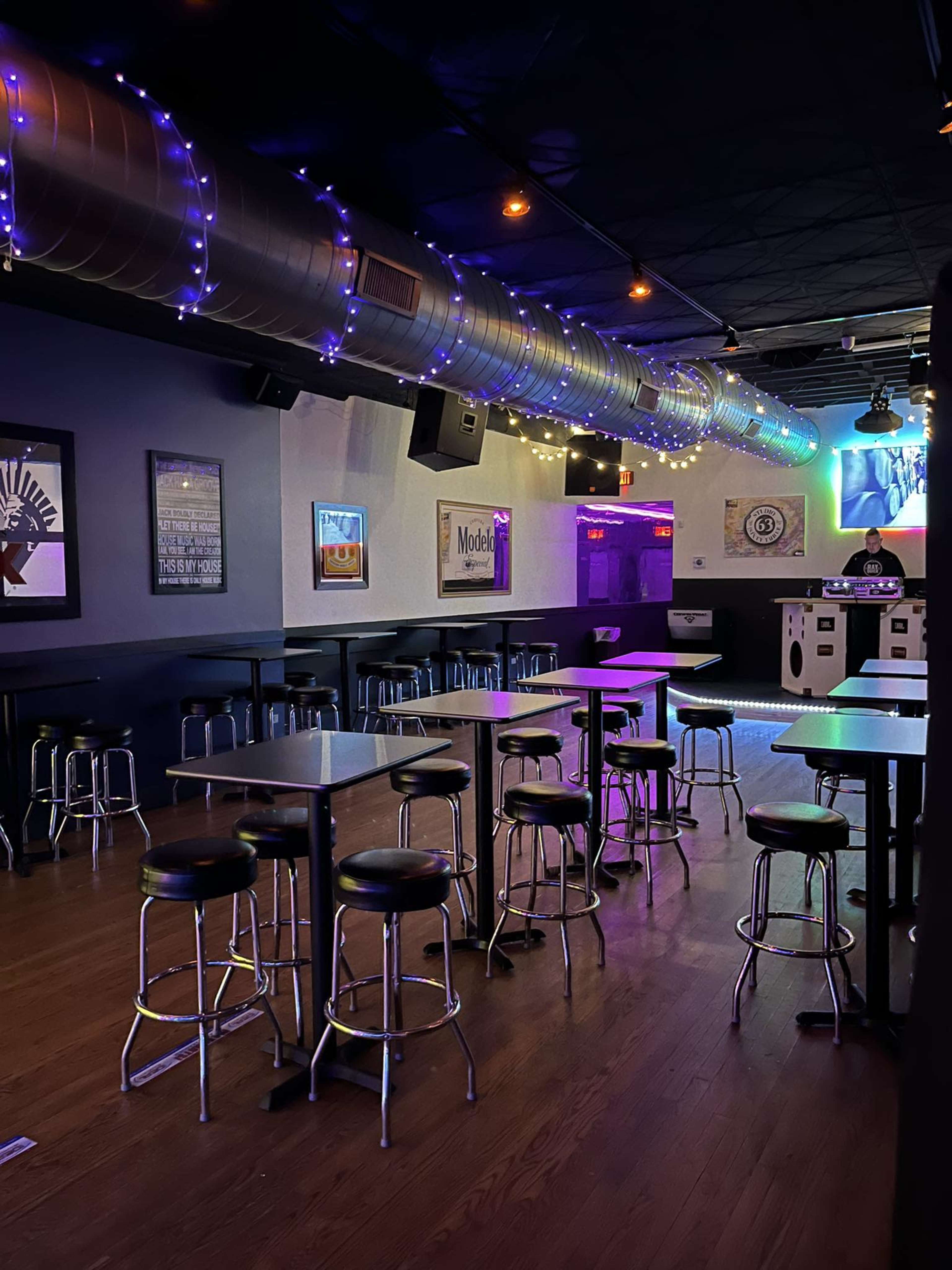 A dimly lit bar features rows of metal stools and tables, with colorful lights illuminating the space and a DJ booth in the background.