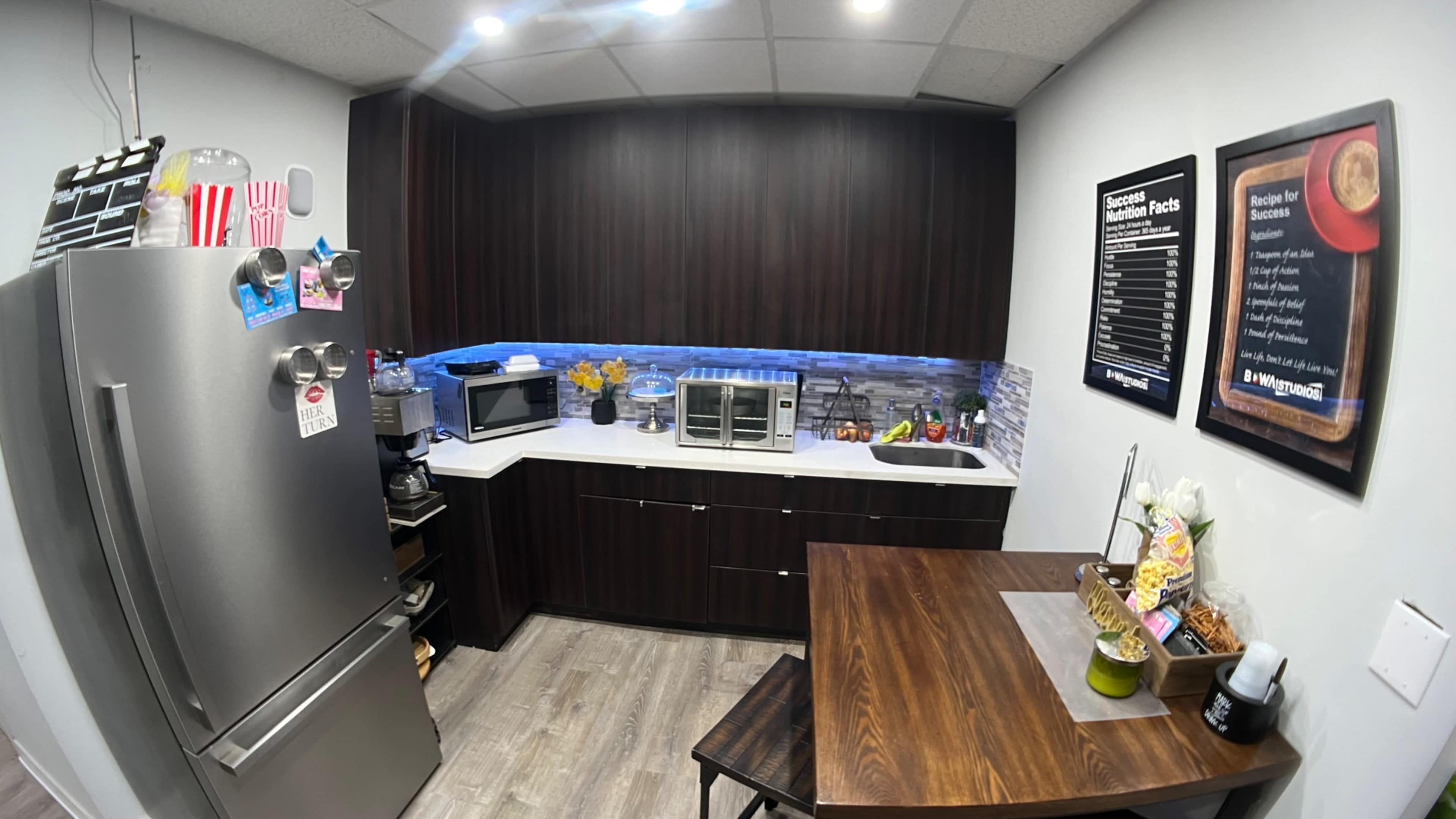 The image shows a modern kitchen with dark cabinetry, a stainless steel refrigerator, a microwave, a toaster oven, and a small dining table.