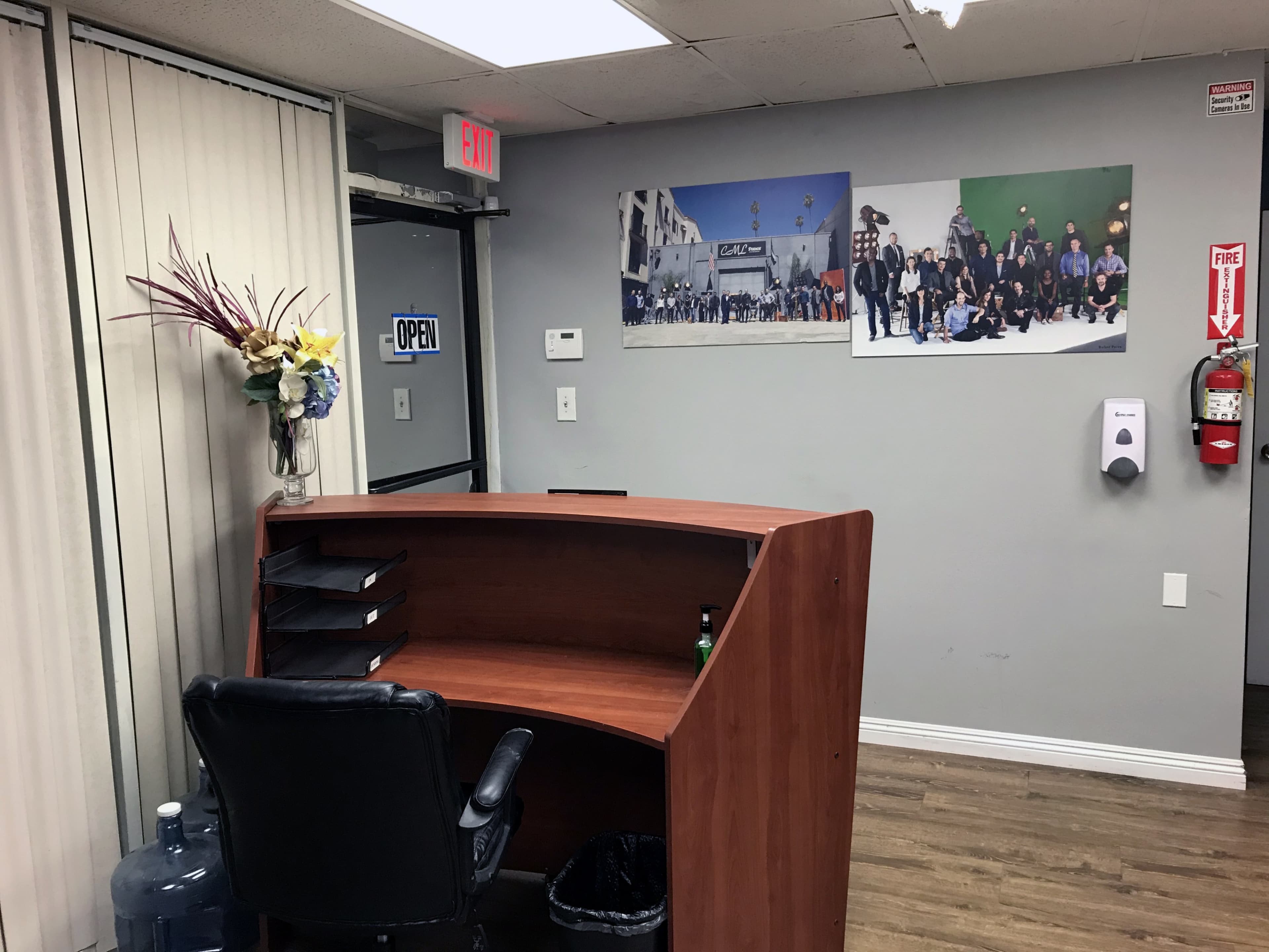 A reception area with a wooden desk, a chair, a water cooler, decorative flowers, an open sign, and a wall displaying a large group photo.