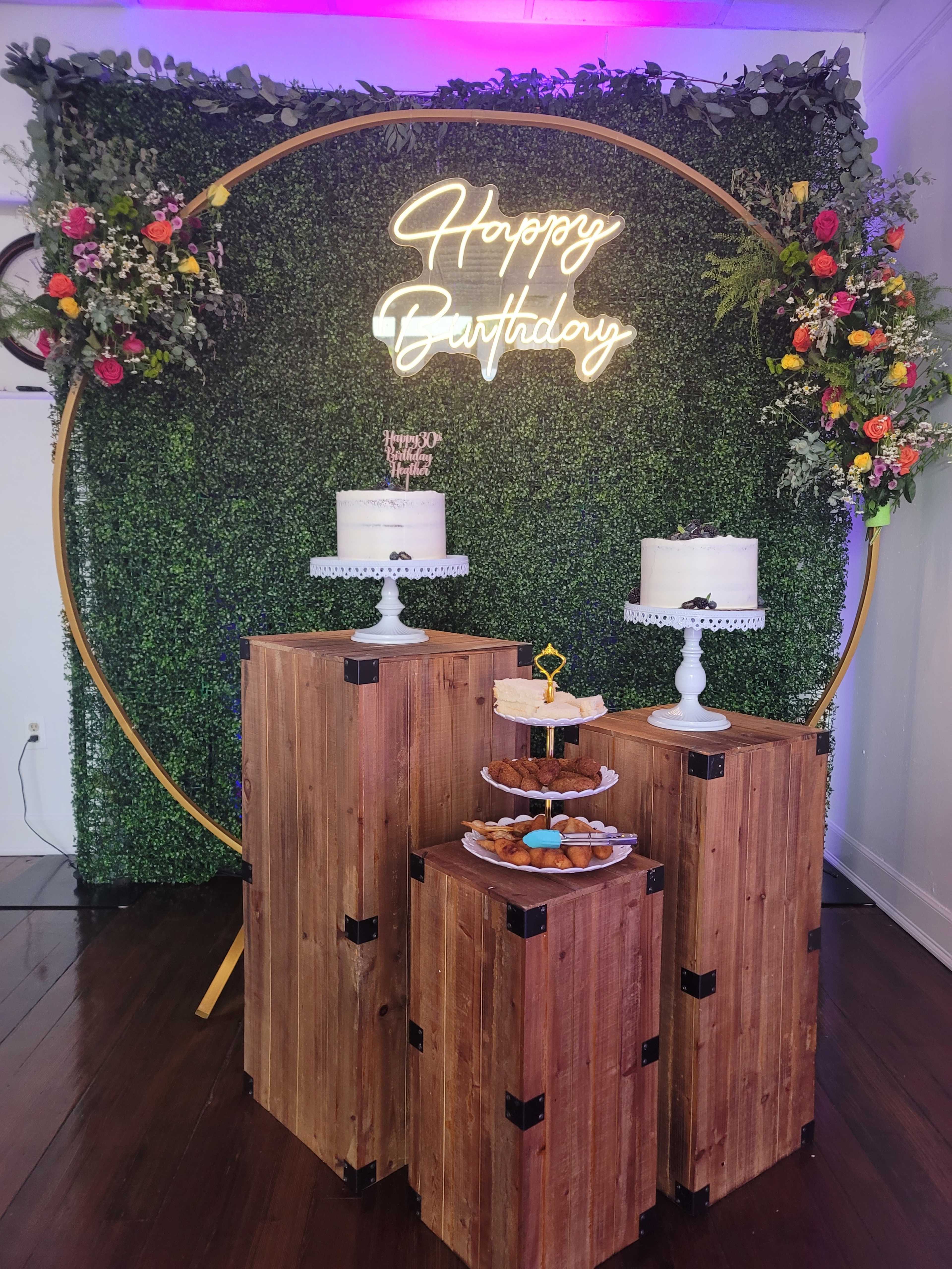 The image features a birthday celebration setup with a neon "Happy Birthday" sign, two tiered cakes on pedestals, and a dessert display on wooden crates against a green backdrop.