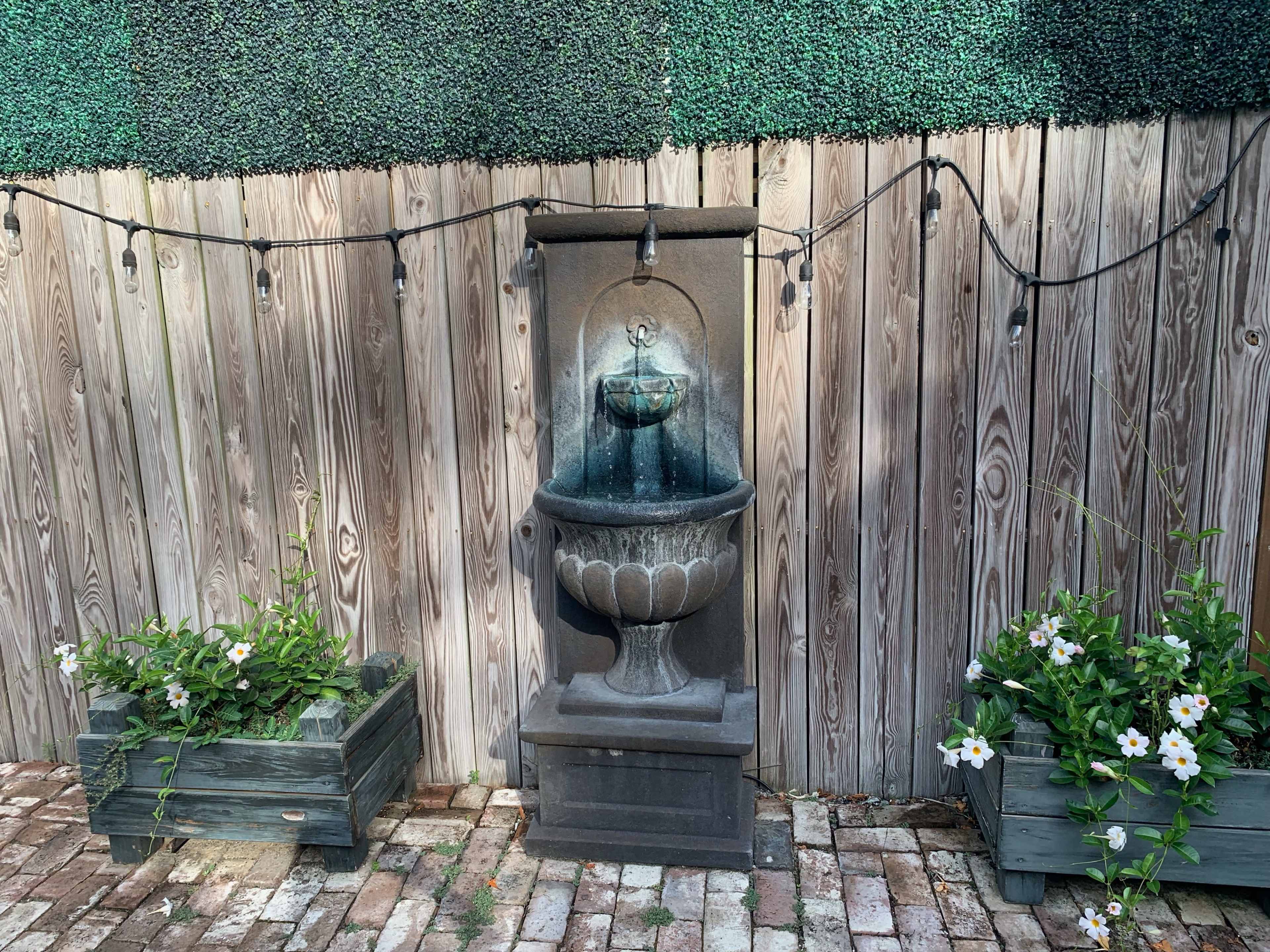 A stone fountain is positioned against a wooden fence, surrounded by two planter boxes filled with flowers and strung with decorative lights.