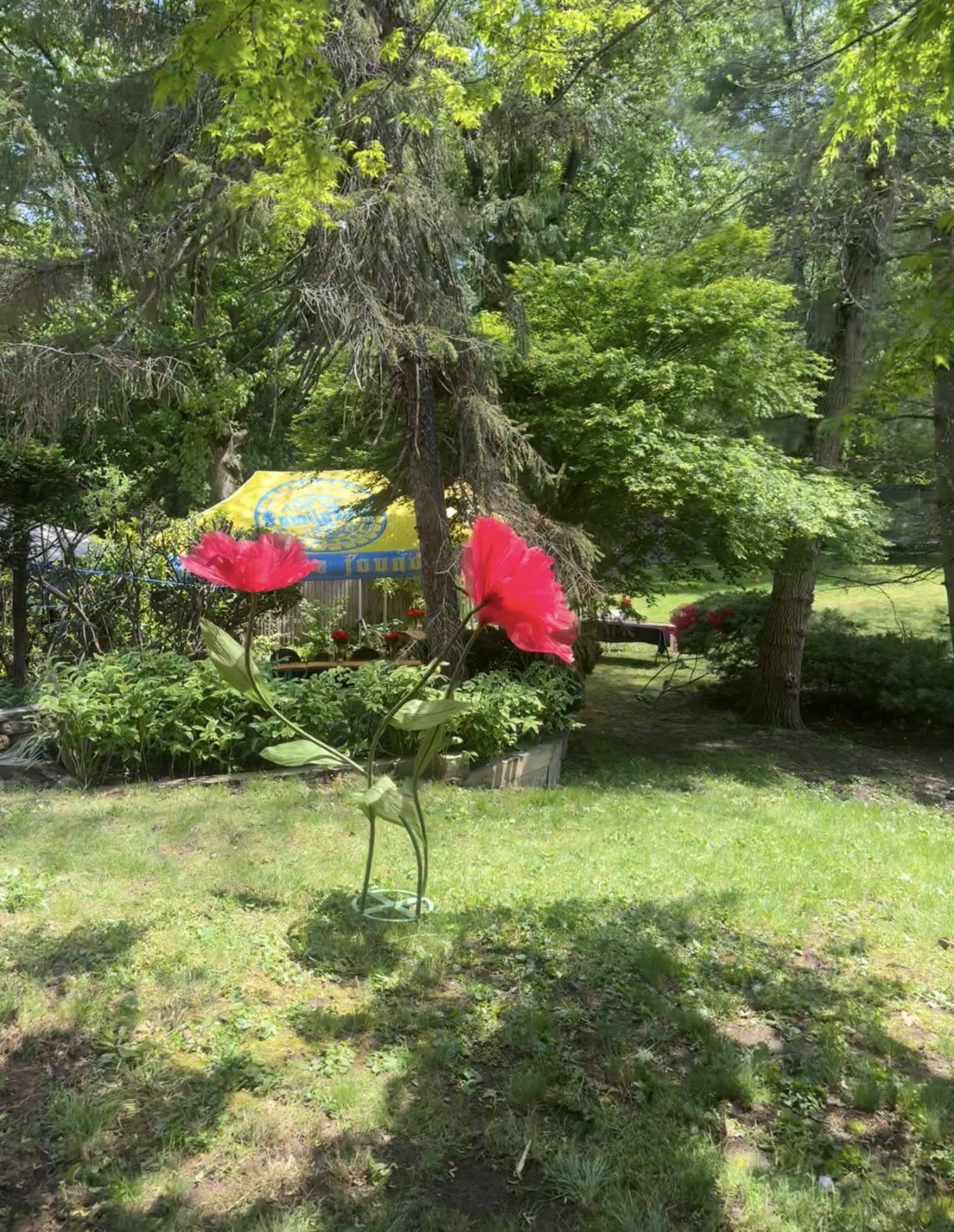 A large metal flower sculpture with red petals stands in a green yard surrounded by trees and a colorful tent in the background.