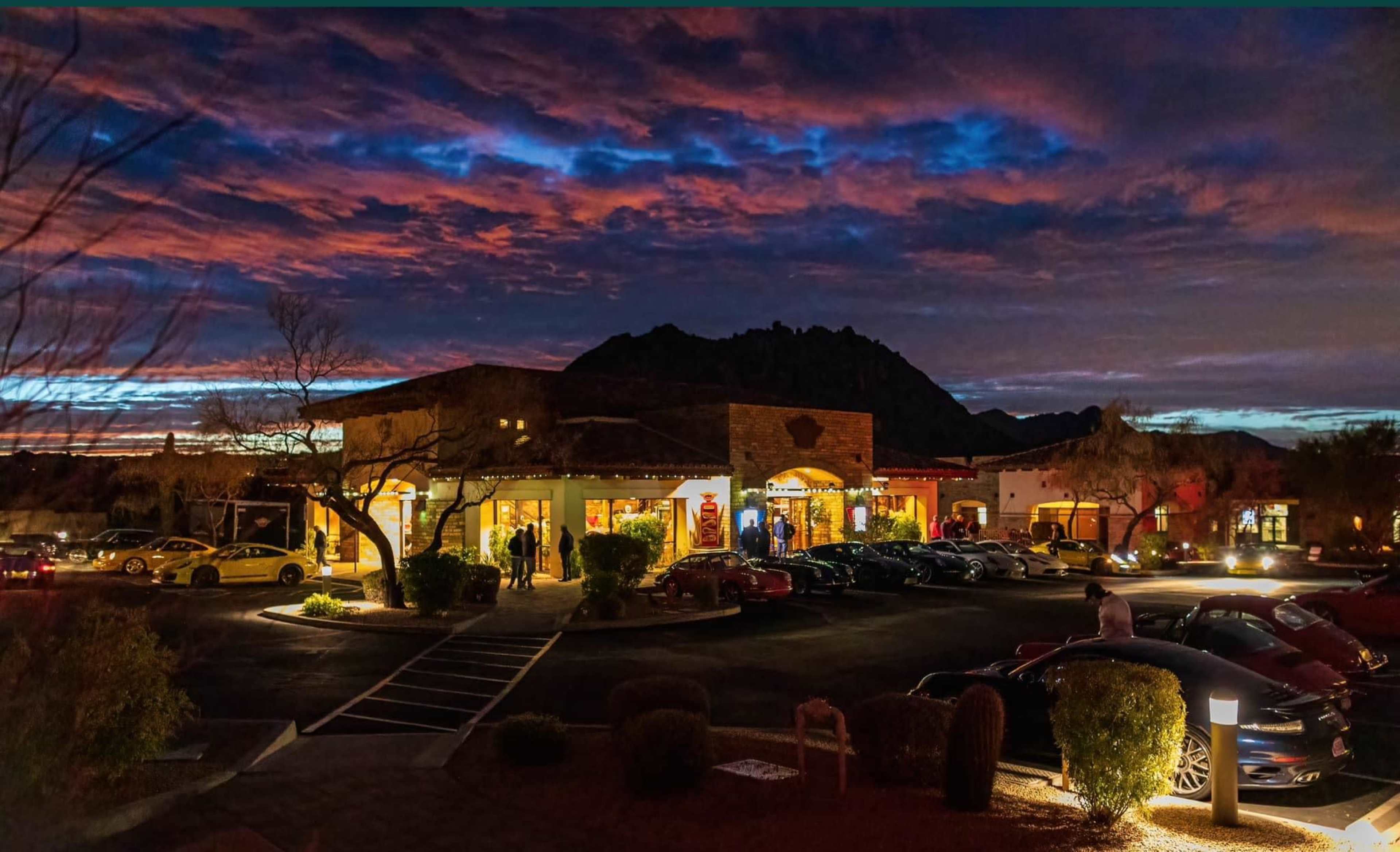 A commercial building with outdoor seating and several parked cars is illuminated under a colorful sunset sky and surrounded by mountains.